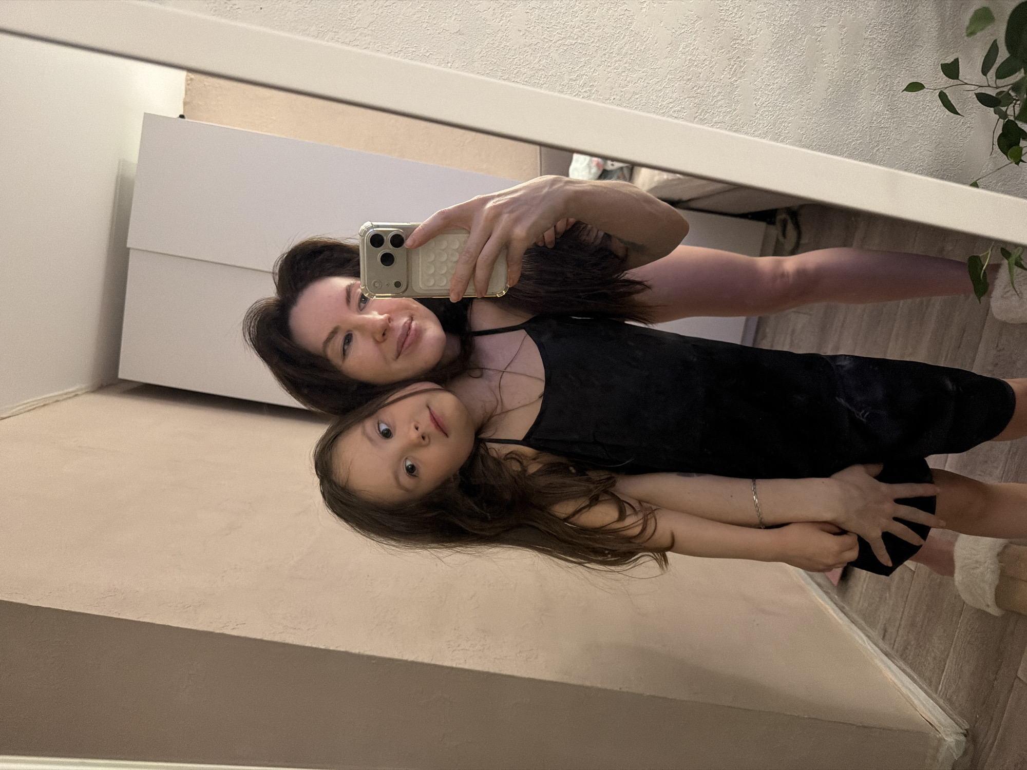 An indoor mirror selfie captures two females, likely a mother and daughter, posing together. The image, originally rotated 90 degrees clockwise, shows them reflected in a large mirror.
The adult woman, positioned behind the young girl, has dark brown hair and is wearing a black strap top. She holds a smartphone, resembling an iPhone with a clear, textured case, extending her arm to capture their reflection. She looks towards the camera with a gentle smile.
In front of her, the young girl also has dark brown hair and wears a black dress or top. She looks directly at the camera with a calm, pleasant expression, her arm wrapped around the woman's waist or hip, suggesting affection.
The setting appears to be a modern home, possibly in Perm, Russia. The background reflected in the mirror includes white furniture, possibly a wardrobe or cabinet, to the left. The walls are a light beige or cream color with a textured finish. The flooring is made of wooden planks or laminate. On the right side of the frame, a potted plant with green leaves is partially visible, adding a touch of nature to the indoor scene. The lighting is soft and even, highlighting their faces and the clean aesthetic of the room. No visible text is discernible in the image.