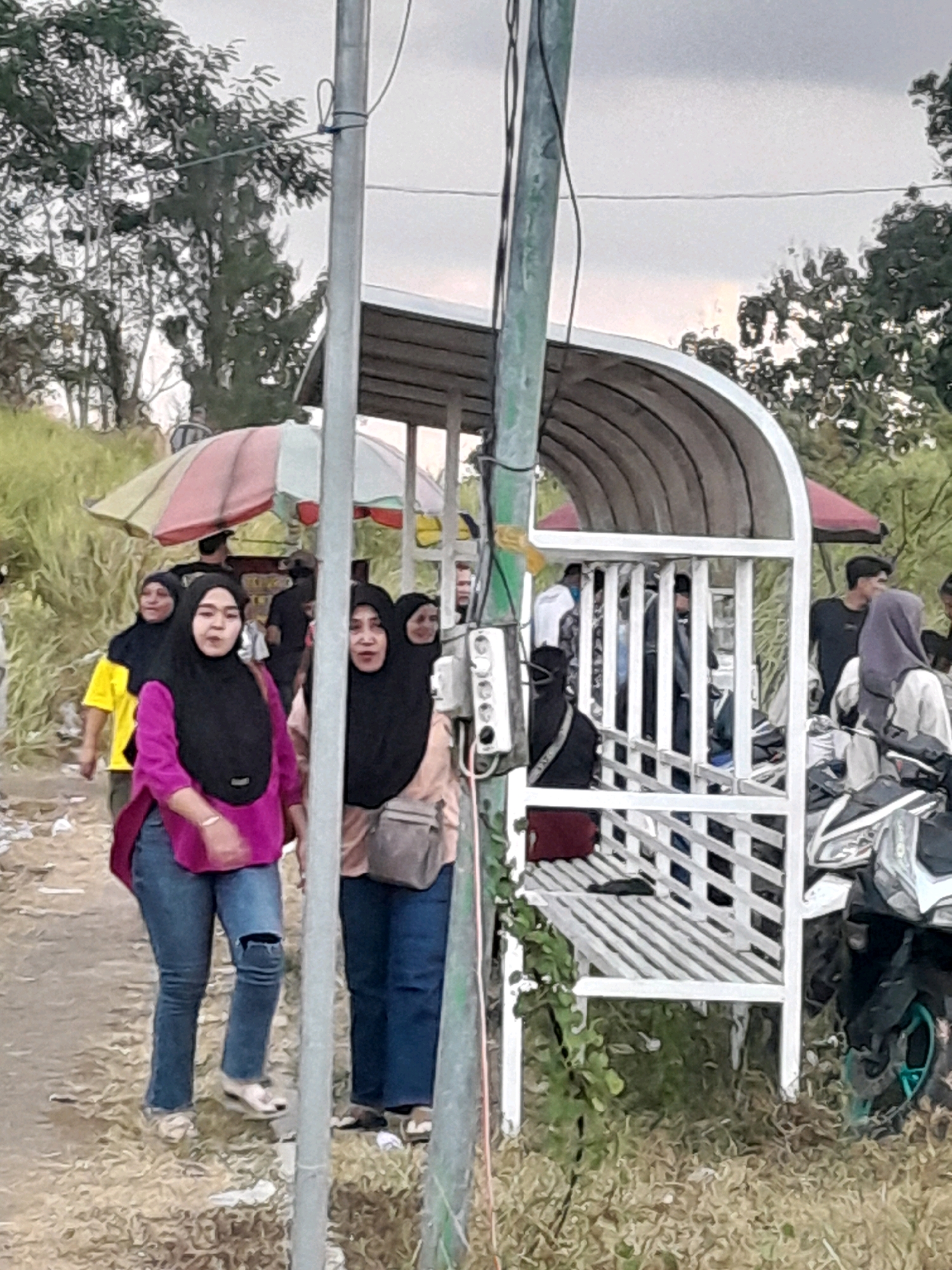 In Genteng, Indonesia, a group of people are gathered near a covered seating area with a white latticework front. Two women in hijabs are in the foreground, one wearing a magenta top and ripped blue jeans, the other in a pale peach top with a grey crossbody bag. Behind them, another woman in a yellow shirt and black hijab walks away. Several other individuals, some obscured, stand or sit within and around the shelter. A motorcycle is parked to the right of the structure, and a striped umbrella is visible over the seating area. The ground is a mix of dirt and dry vegetation, with trees and foliage in the background under an overcast sky. A utility pole with electrical wires and a junction box stands prominently in the center of the frame.