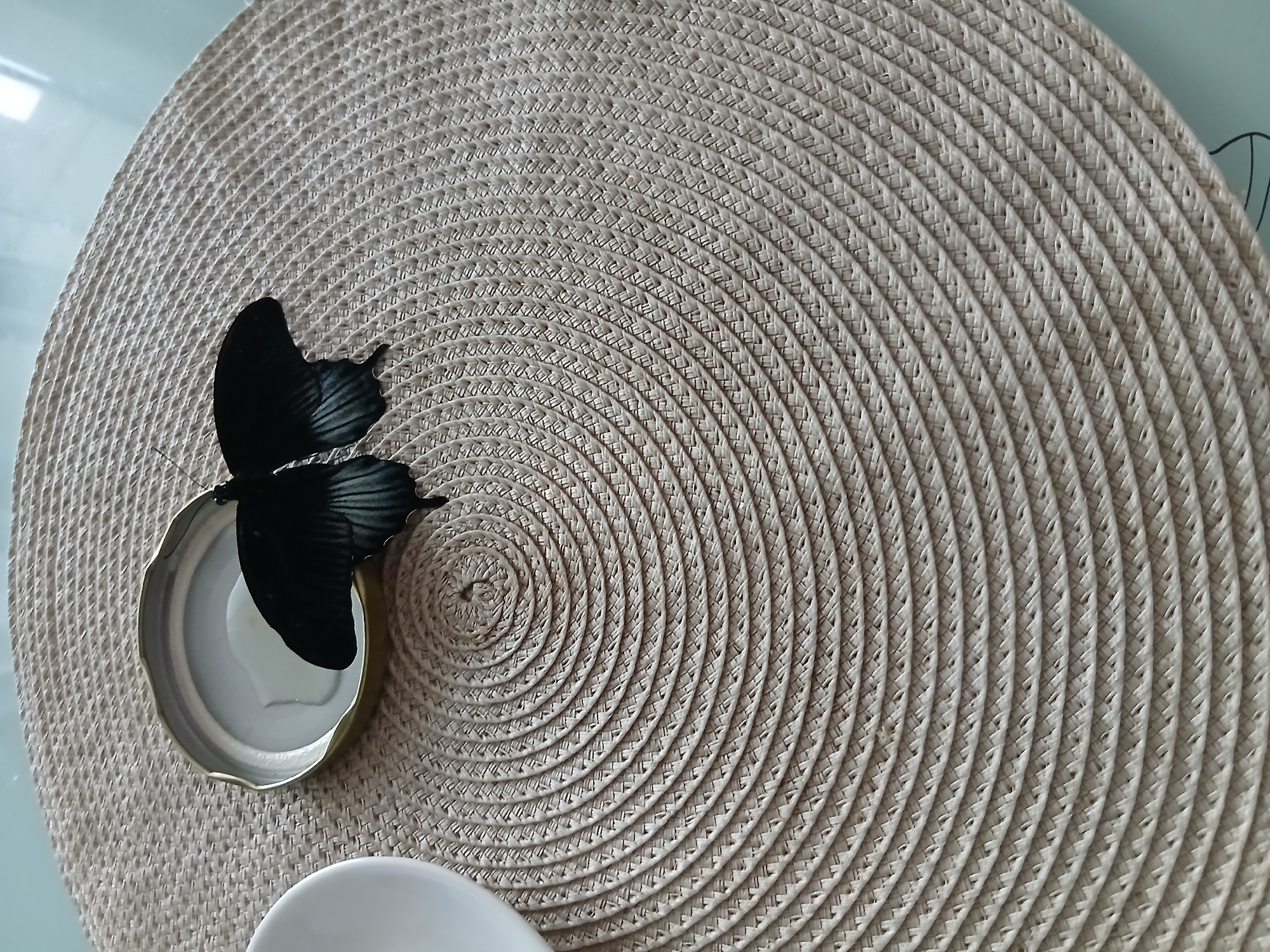A black butterfly is resting on the inner side of a silver jar lid, which contains a small amount of white liquid. The lid is positioned on a light-colored, circular placemat with a woven texture. To the left of the lid, a portion of a white bowl is visible. The placemat is placed on a light-colored, smooth surface, possibly a table. The scene is lit from above and to the right, casting subtle shadows.