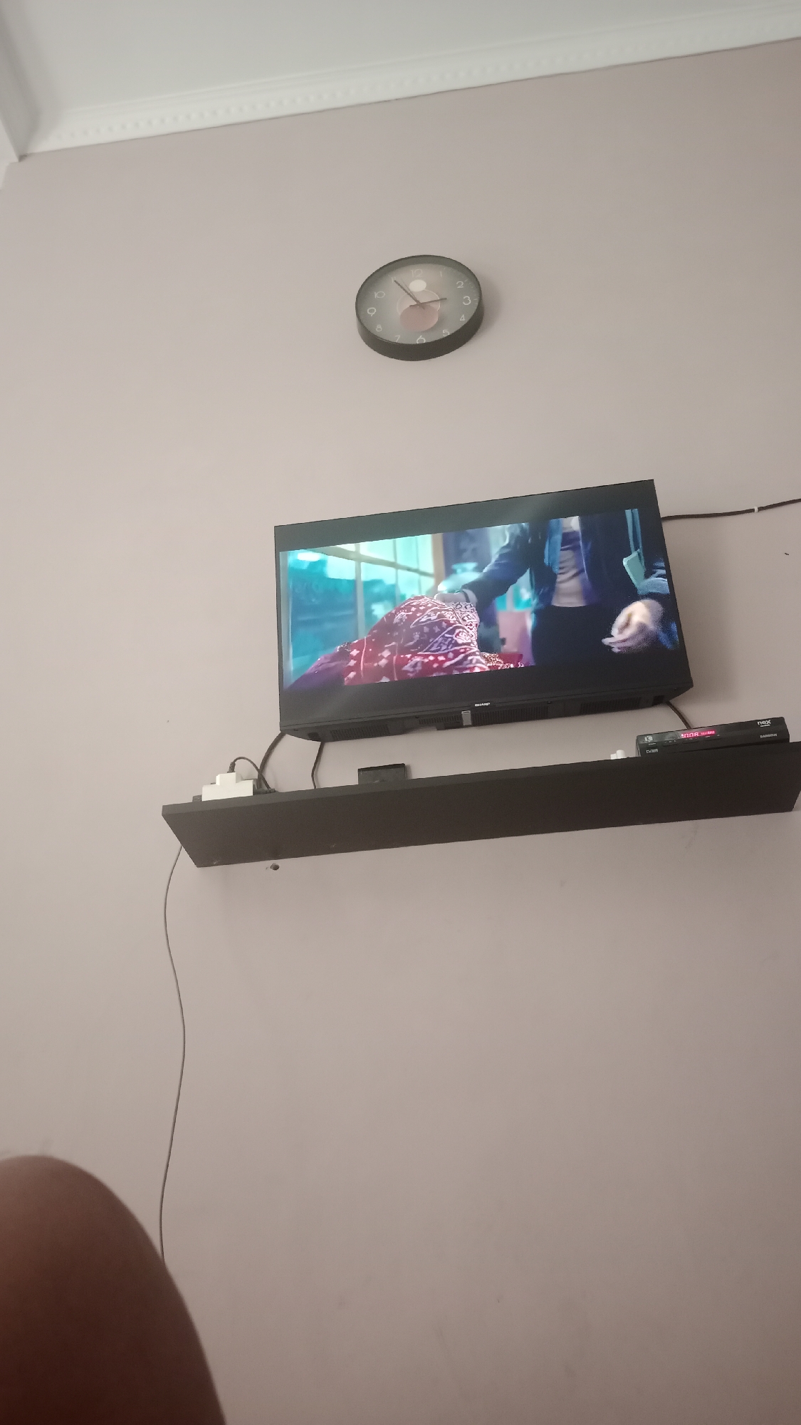 The image shows a television mounted on a shelf against a light pink wall. A round clock with a dark frame and a pink center is visible on the wall above and to the left of the television. The television screen displays a scene from a movie or show, featuring a person in a red and white patterned garment and another person in dark clothing. Below the television, on the shelf, are several electronic devices, including what appears to be a set-top box and a power strip with cables. A thin grey cable hangs down from the shelf. The lower left corner of the image shows a blurry portion of a person's leg or arm. The environment appears to be an indoor setting, likely a living room. The city of Pakemitan, Indonesia is not depicted in this image.