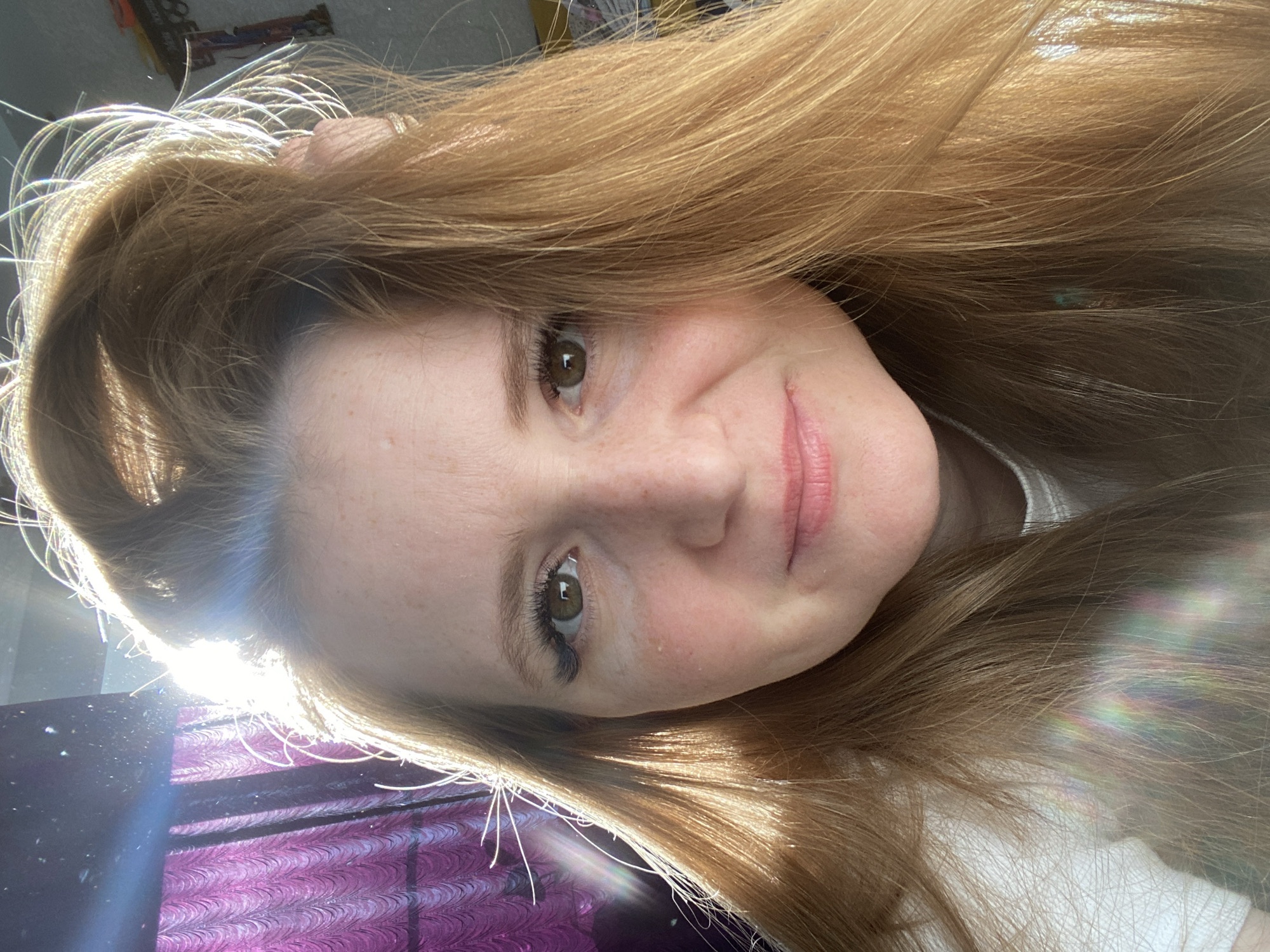 A close-up, rotated selfie depicts a woman with light brown hair and fair skin. Her face is angled towards the camera, and she's smiling slightly. Her eyes are light brown. The background is out of focus but reveals a purple and black surface, likely a curtain or decorative cloth. Bright sunlight illuminates the lower left corner of the image. No information indicates that the image was taken in Chaikovsky, Russia.