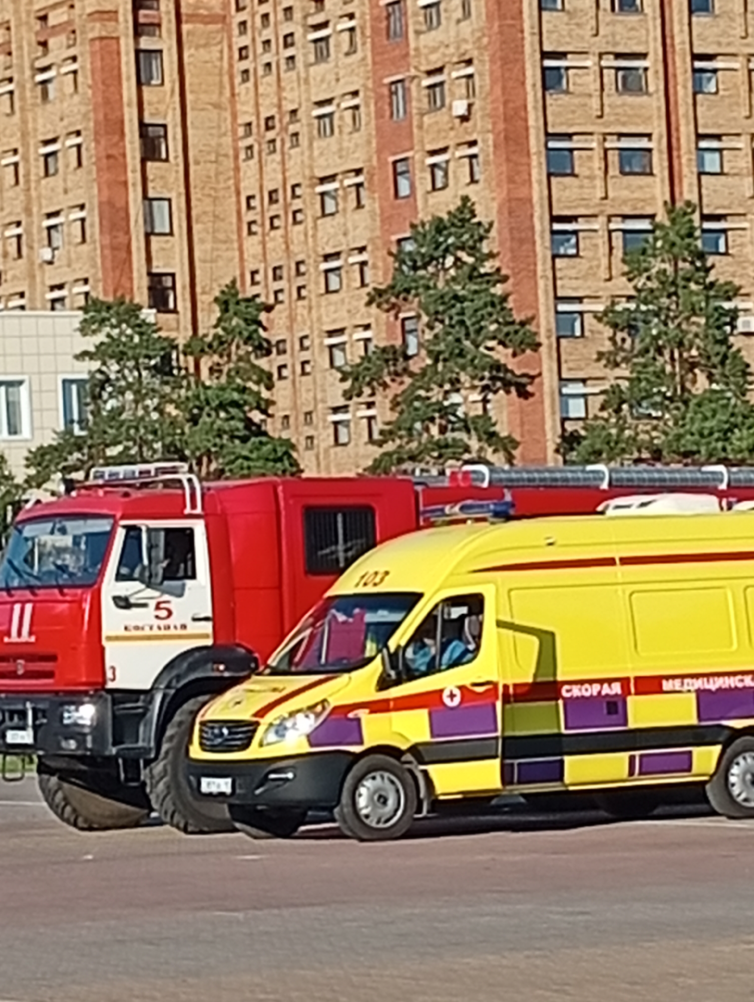 In the city of Kostanay, Kazakhstan, a bright red fire truck is parked alongside a yellow ambulance. The fire truck, marked with the number "5" and "KOCTAHAD" (Kostanay in Cyrillic), is positioned to the left of the ambulance. The ambulance, also displaying the word "СКОРАЯ" (Emergency) and "МЕДИЦИНСКАЯ" (Medical) in Cyrillic, features a distinctive red and purple checkered stripe. The vehicles are situated on a paved surface, likely a street or parking lot, with a large, brick building forming the backdrop. Trees with green foliage are visible between the vehicles and the building, suggesting an outdoor setting. The lighting indicates it is daytime, with sunlight illuminating the scene. There are no visible people interacting with the vehicles in the provided image.