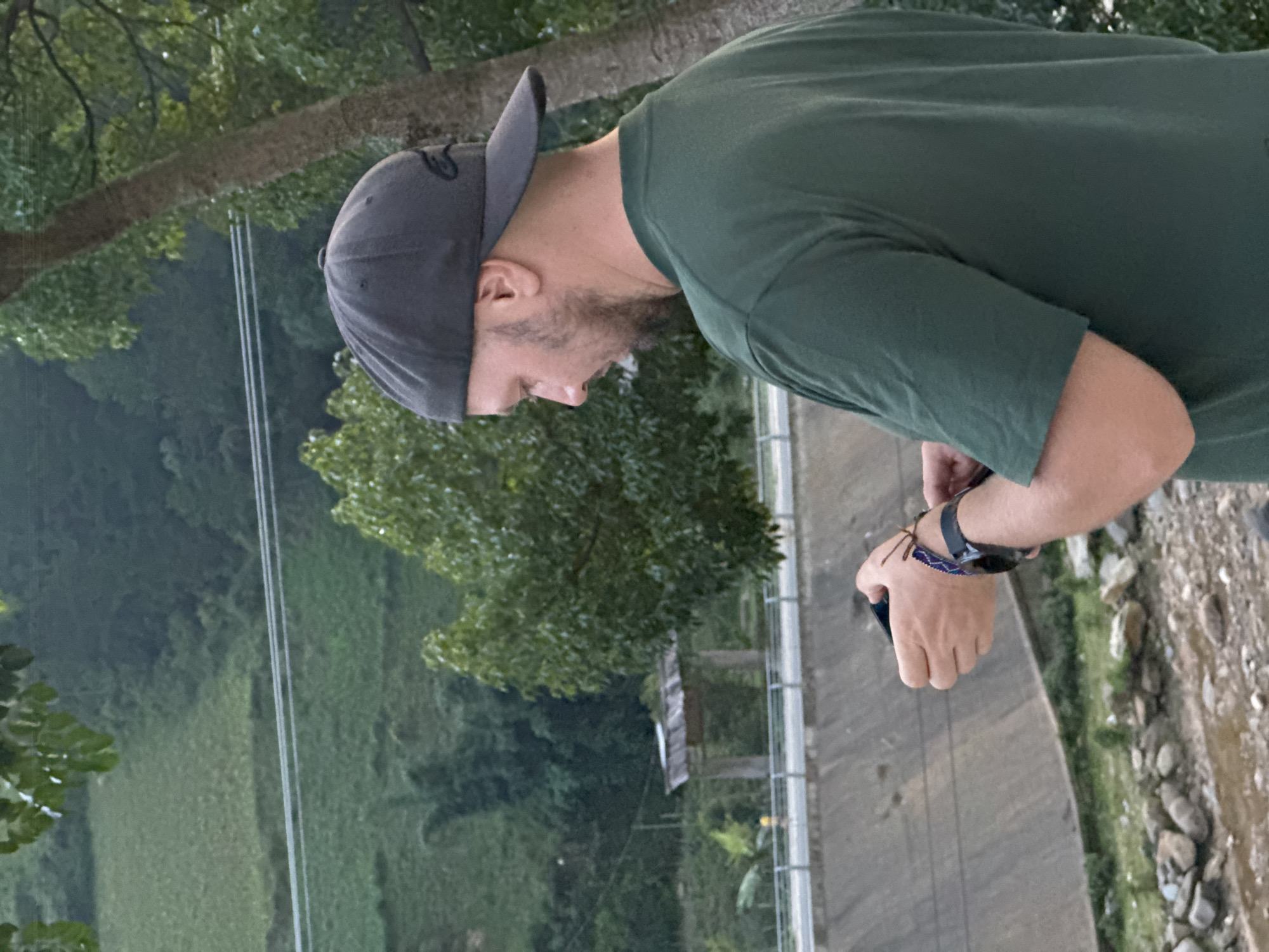 A man in a dark green t-shirt and a dark grey baseball cap stands outdoors, viewed from his left side, looking downwards. He has a beard and is holding a dark object, possibly a smartphone, in his hands. On his left wrist, he wears a watch and a patterned, braided bracelet.
He is positioned on a paved or concrete path, next to a low metal fence or railing. Beyond the railing, a lush, verdant valley or slope stretches into the background, densely covered with green foliage and trees. The landscape suggests the terraced fields characteristic of Mù Cang Chải, Vietnam. Utility lines are visible spanning across the mid-ground. To the man's right, below the path, there is a rocky, dry gully or riverbed with sparse vegetation.
The lighting is soft and diffused, indicating either an overcast day or the late afternoon/early evening hours. The man appears focused on the object in his hands. No visible text is discernable on the man's clothing or in the surrounding environment.