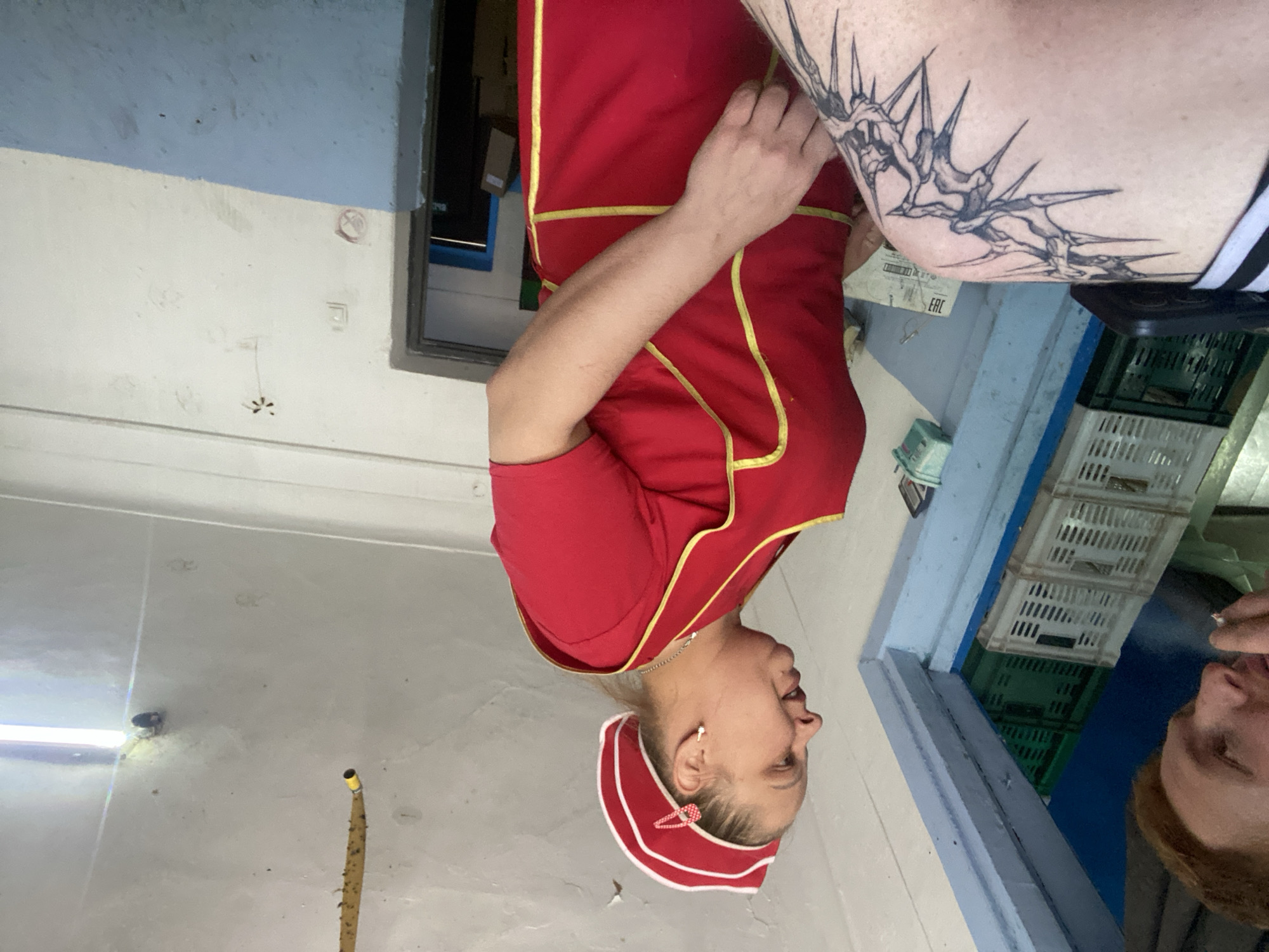 A man with red hair is looking down, holding a cigarette, and talking to a woman wearing a red uniform with yellow trim and a matching hat. The woman is on the right side of the frame, facing left. Behind her is a doorway leading to another room. In the background, there are stacks of white and green plastic crates. The floor is tiled and there is a blue-painted wall on the left. A tattoo is visible on the lower left side of the frame, on a person's leg or arm. The ceiling is white and shows some signs of wear, with a yellow object hanging down.