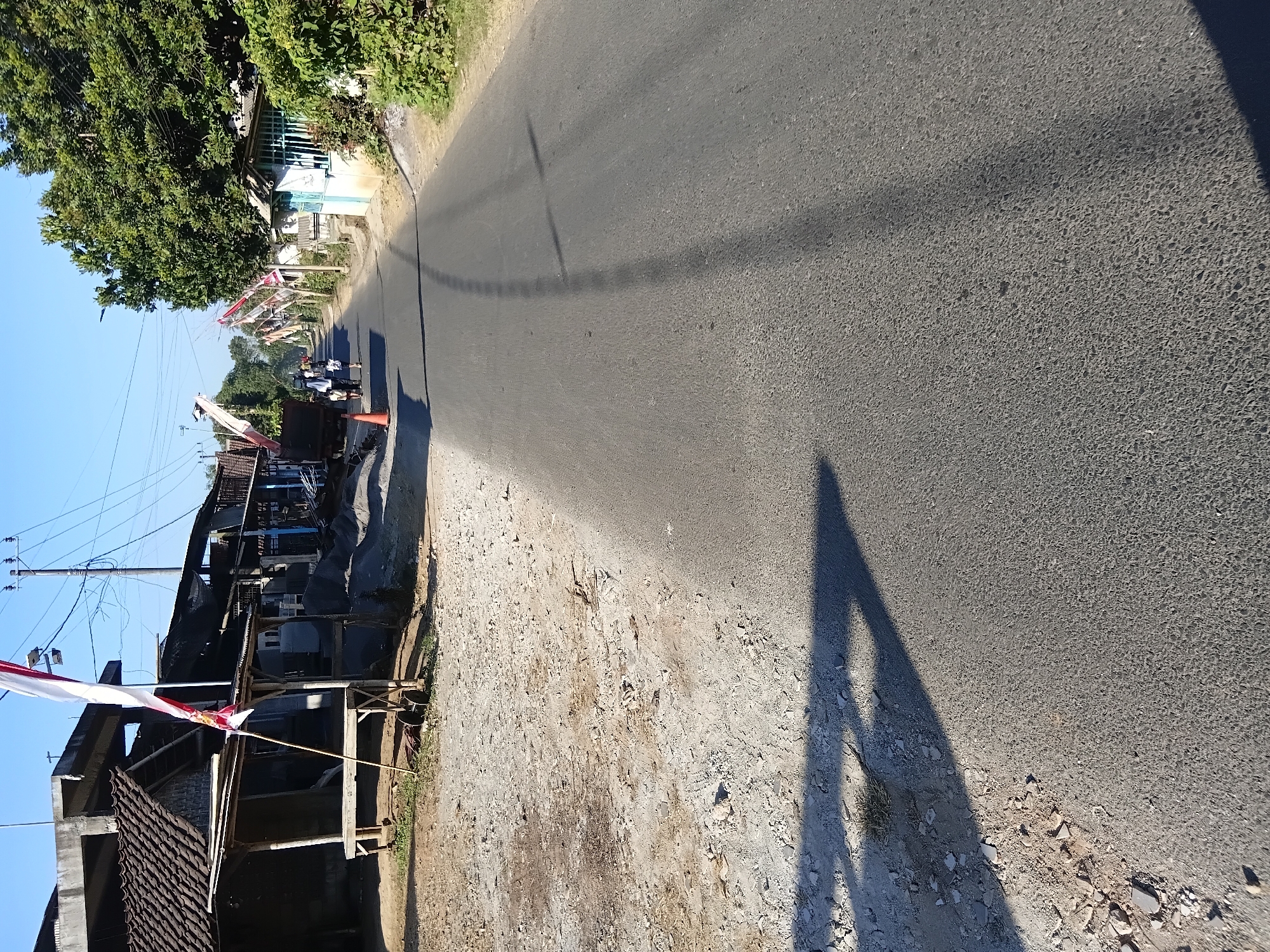The image depicts a roadside scene in Tanggul Wetan, Indonesia, on a clear day. The road, made of asphalt, stretches into the distance, with a dusty, unpaved shoulder on the left. Several structures line the right side of the road, including a house with a blue-green fence and lush green vegetation. On the left side, a small wooden stall with a tiled roof is visible, accompanied by a large pile of construction materials. A flagpole with an Indonesian flag stands prominently near the stall.
In the middle ground, two people are walking away from the camera, down the road. One is wearing a white headscarf and a dark outfit, while the other is wearing a light-colored outfit. A large truck is parked on the left side of the road, near the construction materials. Decorative banners or flags are strung along the right side of the road, suggesting a festive occasion or celebration. The sun casts long shadows across the road and the surrounding ground.
