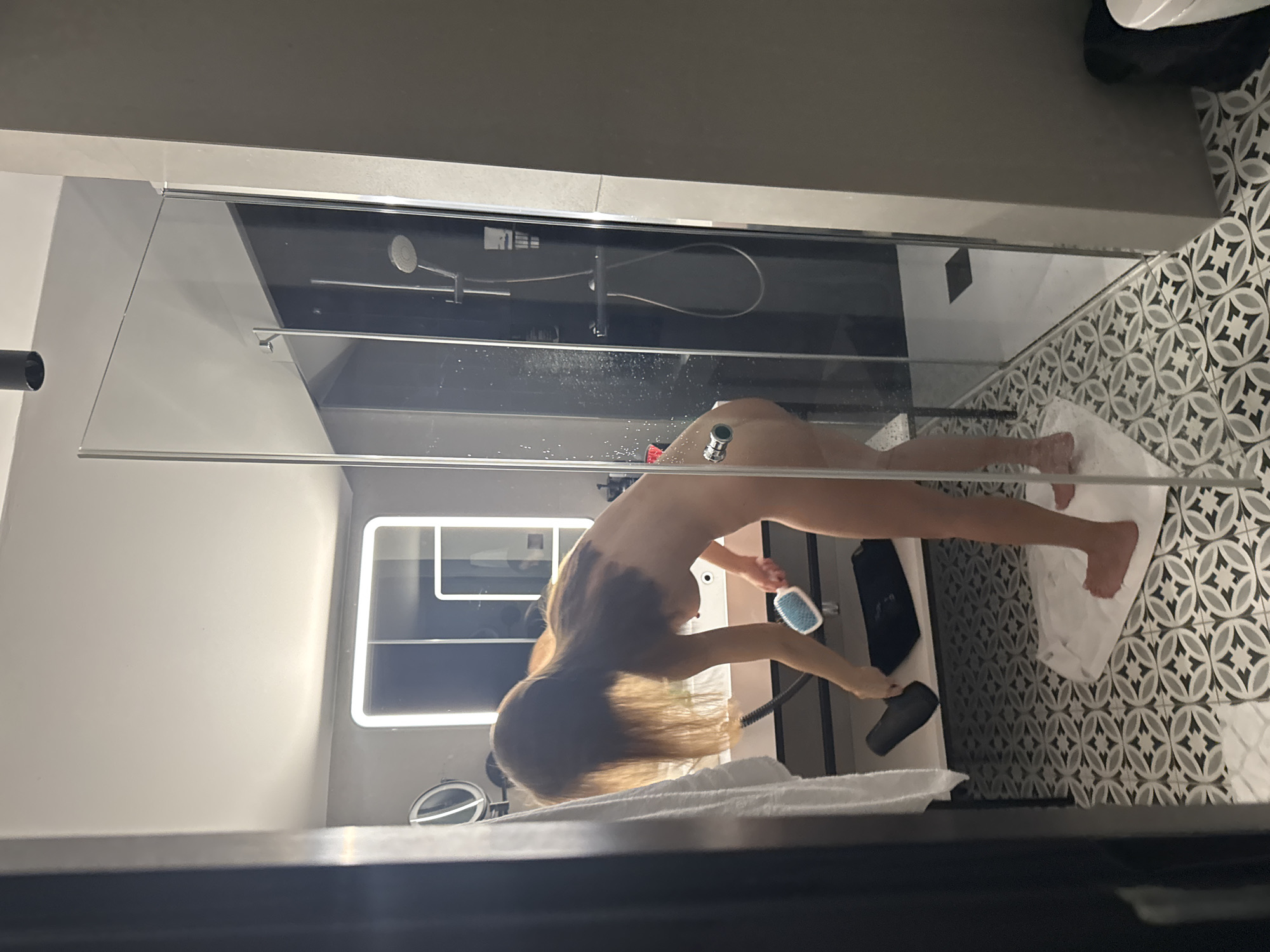 The image shows a nude person inside a modern glass shower enclosure, viewed from outside. The person, with long, light brown hair, is bent over at the waist, allowing their hair to hang down towards the floor. They are holding a blue and white hairbrush in one hand and a black hairdryer in the other, suggesting they are in the process of drying their hair.
The shower enclosure features clear glass panels and a metallic frame. A shower head and hose are mounted on the back wall of the shower. Water droplets are visible on the interior surface of the glass door, indicating recent use of the shower. The person is standing on a white bath mat or towel placed on the patterned floor inside the enclosure. The bathroom floor outside the shower is covered with decorative black, white, and grey geometric tiles.
In the background, through the shower glass, a contemporary bathroom setting is visible. This includes a large wall-mounted mirror with an illuminated edge and a counter with a smaller, circular vanity mirror. A white towel is partially visible in the foreground, draped over a surface. The scene is well-lit by indoor artificial lighting. No discernible text is visible in the image.