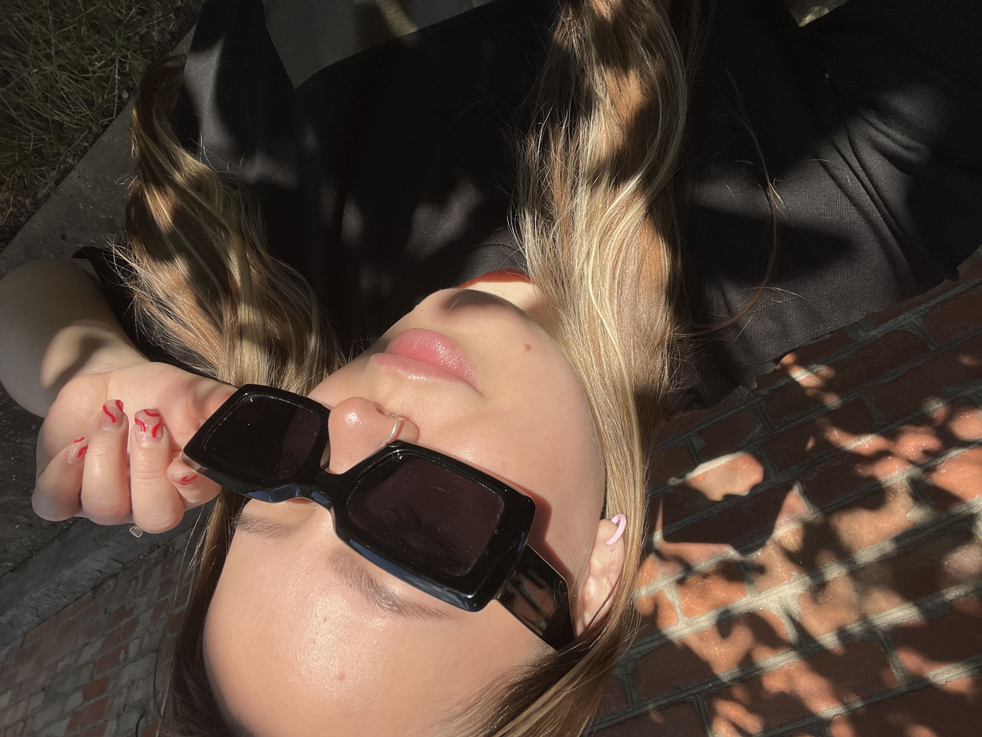 The media file shows a close-up, overhead shot of a person lying down outdoors in Novopokrovskaya, Russia. The subject is wearing black sunglasses and a black top. Their blonde hair, with lighter streaks, is spread out, catching the sunlight. A hand with painted red nails is raised near the sunglasses. A light pink earring is visible on one ear. The background consists of a brick wall and some grassy area, with dappled sunlight and shadows cast by foliage, suggesting a sunny daytime. The overall mood appears relaxed and summery. No visible text is present in the media file.