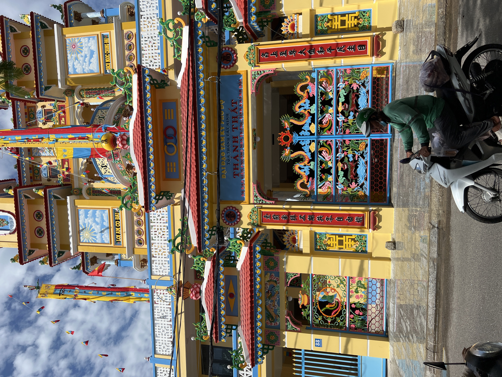 This image depicts a vibrant yellow building with red tiled roofs, characteristic of Vietnamese temple architecture. The building is adorned with intricate dragon motifs, colorful banners, and what appear to be religious inscriptions in Vietnamese. A tall, multi-tiered structure rises in the background, topped with what looks like a golden orb and religious symbols. Small triangular flags are strung across the sky. In the foreground, a person wearing a green long-sleeved shirt, dark pants, and a green helmet is riding a white motorcycle with a bag secured to the back. The motorcycle is on a paved road, and the building is situated behind a sidewalk or pathway. The scene is illuminated by sunlight, creating strong shadows. The building's signage indicates it is "ĐẠI ĐẠO TAM KỲ PHỔ ĐỘ - THÁNH THẤT", suggesting it is a Caodaist temple. The presence of "NHA TRANG, KHÁNH HÒA" on the signage confirms the location as Nha Trang, Vietnam.