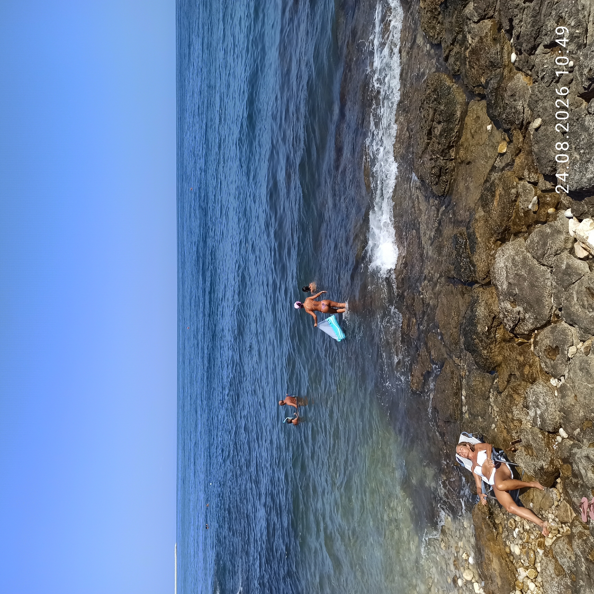 The image depicts a sunny day at a rocky coastline, likely in Molfetta, Italy, as indicated by the superimposed date "24.08.2026 10:49". The scene shows several people enjoying the water and beach area.
In the foreground, a woman lies on a white beach chair on the rocky shore, wearing a white bikini. Her eyes are closed, suggesting she is sunbathing or relaxing. Nearby, a pair of pink flip-flops rests on the rocks.
Further into the water, a woman in a patterned bikini is wading out, carrying a blue and white inflatable mat. Another person, partially obscured, is walking in the water behind her. Further out, a man and a child are in the sea, with the man wearing a snorkel mask. In the distance, other individuals are visible in the water, some swimming.
The sea is calm with gentle waves lapping at the shore. The coastline is characterized by large, dark rocks and scattered white pebbles. The water transitions from clear near the shore to a deeper blue further out. The sky is a clear, bright blue, indicating fair weather. The presence of a breakwater or pier structure is visible on the horizon, suggesting a developed coastal area.