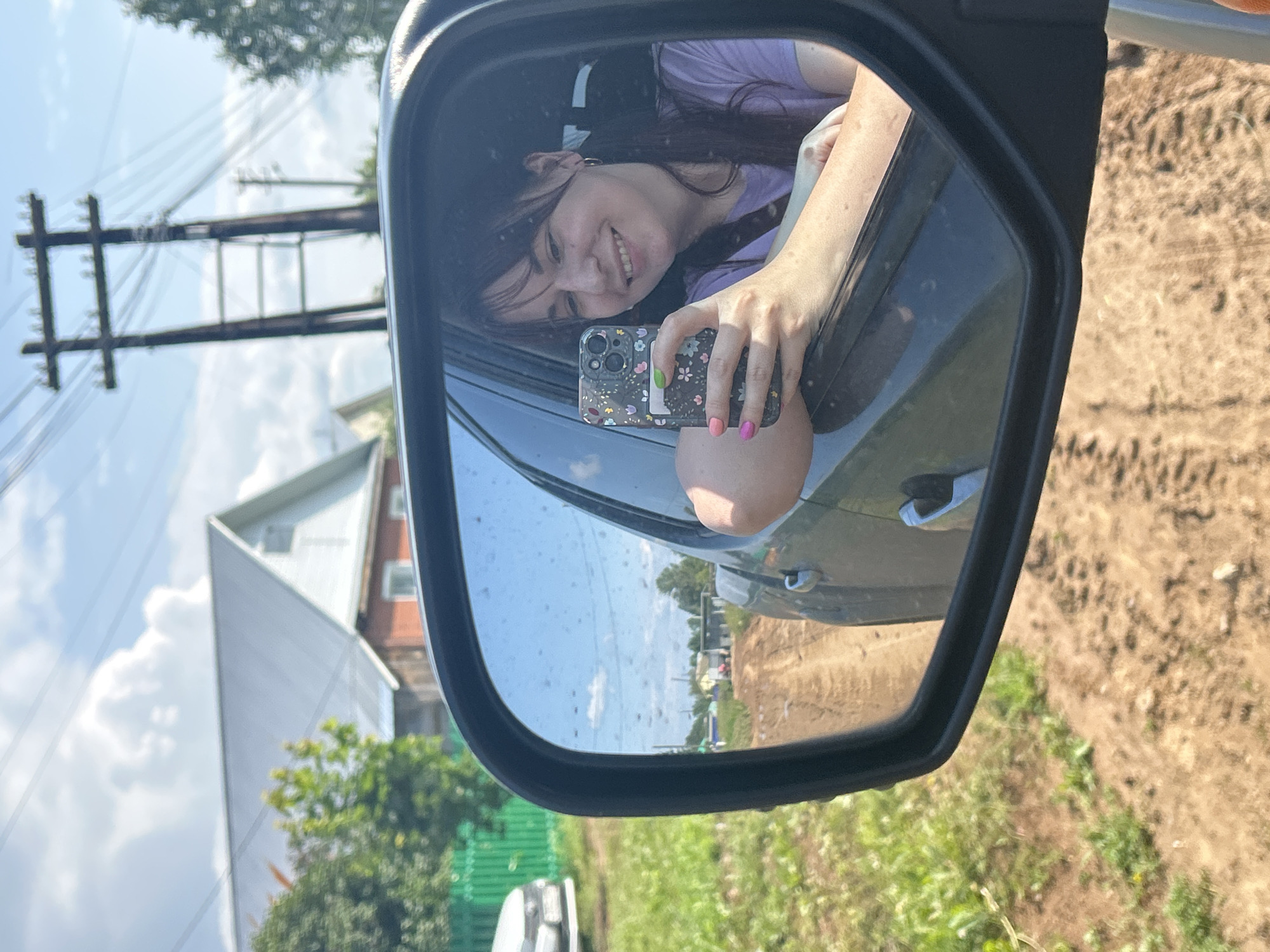 The image is a selfie taken by a young woman in a car, reflected in the side-view mirror. She is smiling and holding a smartphone. Her left arm is resting on the open car door. The car appears to be on a dirt road in a rural area. In the background, there are houses, trees, and power lines. The location is identified as Foki, Russia.