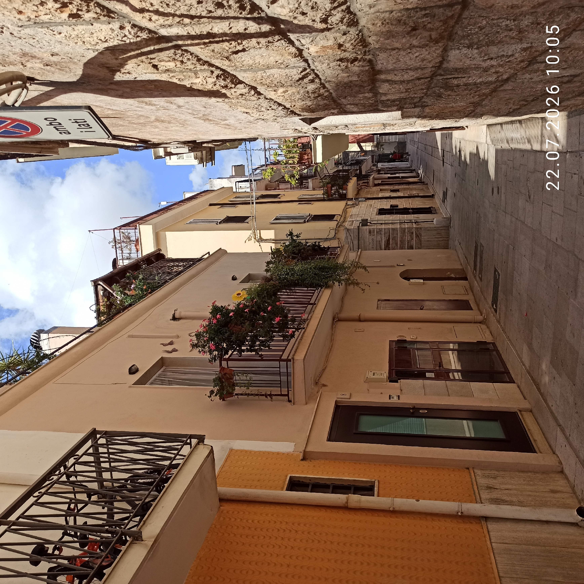 The image depicts a narrow street in Modugno, Italy, likely a residential area. The street is paved with cobblestones and flanked by buildings on both sides. The building on the left has an orange textured facade with a white downspout. It features a dark-framed window and a dark door with a glass panel. Further along, a cream-colored building has balconies adorned with potted plants and flowers, including pink blossoms. The building on the right is constructed with rough-hewn stone blocks and features a traffic sign. The sky above is blue with scattered clouds. The overall scene suggests a quiet afternoon in an Italian town.