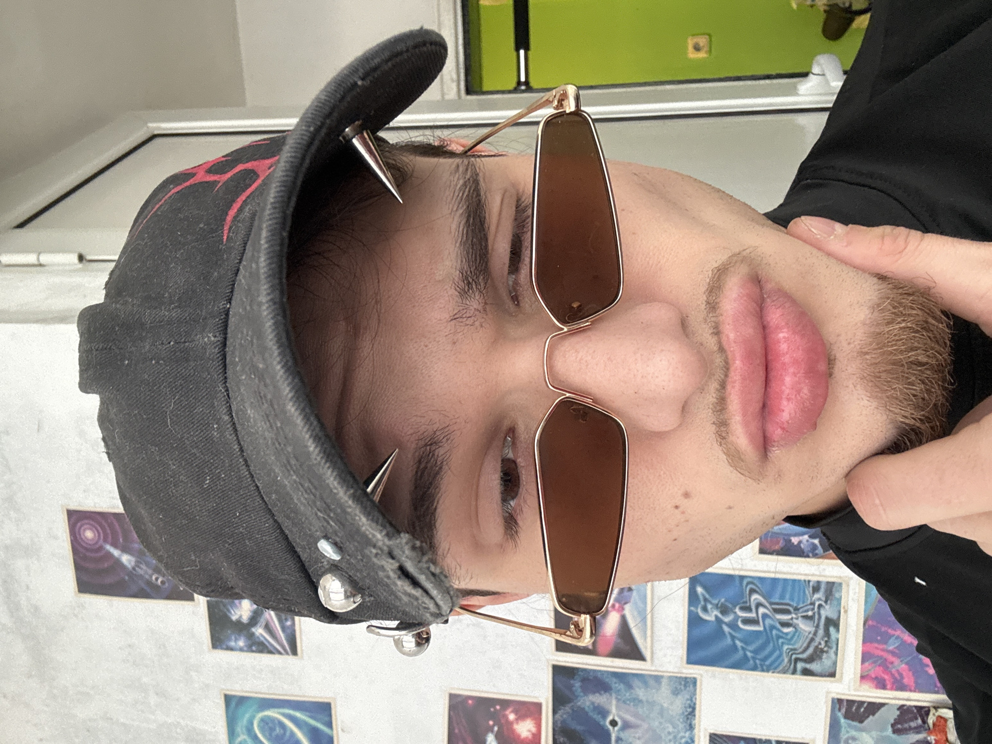 A close-up shot focuses on a person wearing a black cap adorned with metal spikes and studs. The cap also features a red design. The individual is wearing sunglasses with gold frames and brown lenses. Their face is partially obscured by the sunglasses and the cap. The background consists of a white wall with several small framed images or cards. The wall also has a green section at the top. The person is wearing a black shirt or jacket. The location cannot be confirmed based on the image alone; Sofia, Bulgaria cannot be determined from the image.