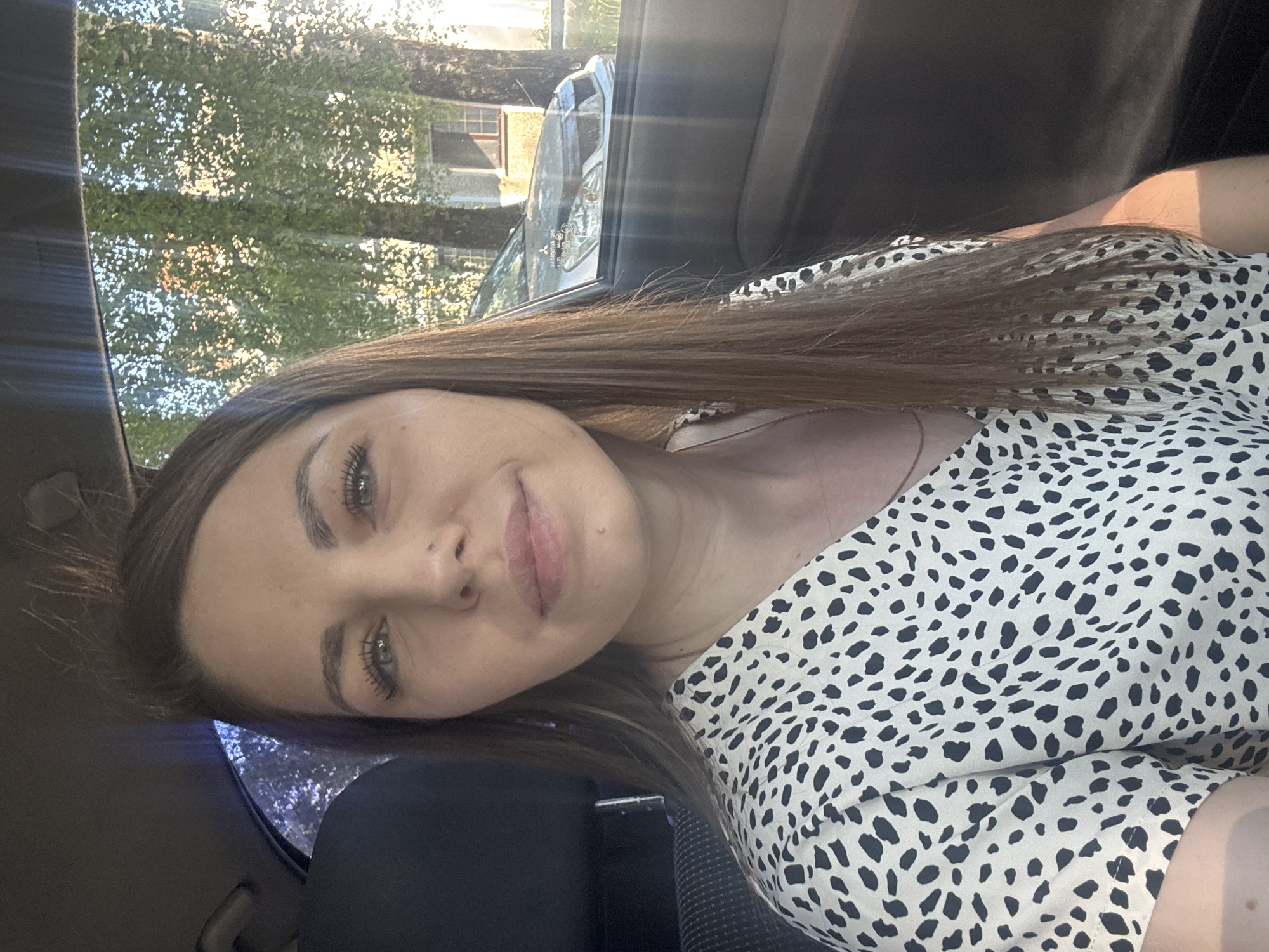 The image presents a woman inside a car. The environment visible through the car window includes trees and a building, suggestive of a residential or suburban area. The interior of the car is dark, focusing attention on the woman's face and upper torso. She is wearing a patterned top. The location is potentially Раздолье, Russia based on user instructions.