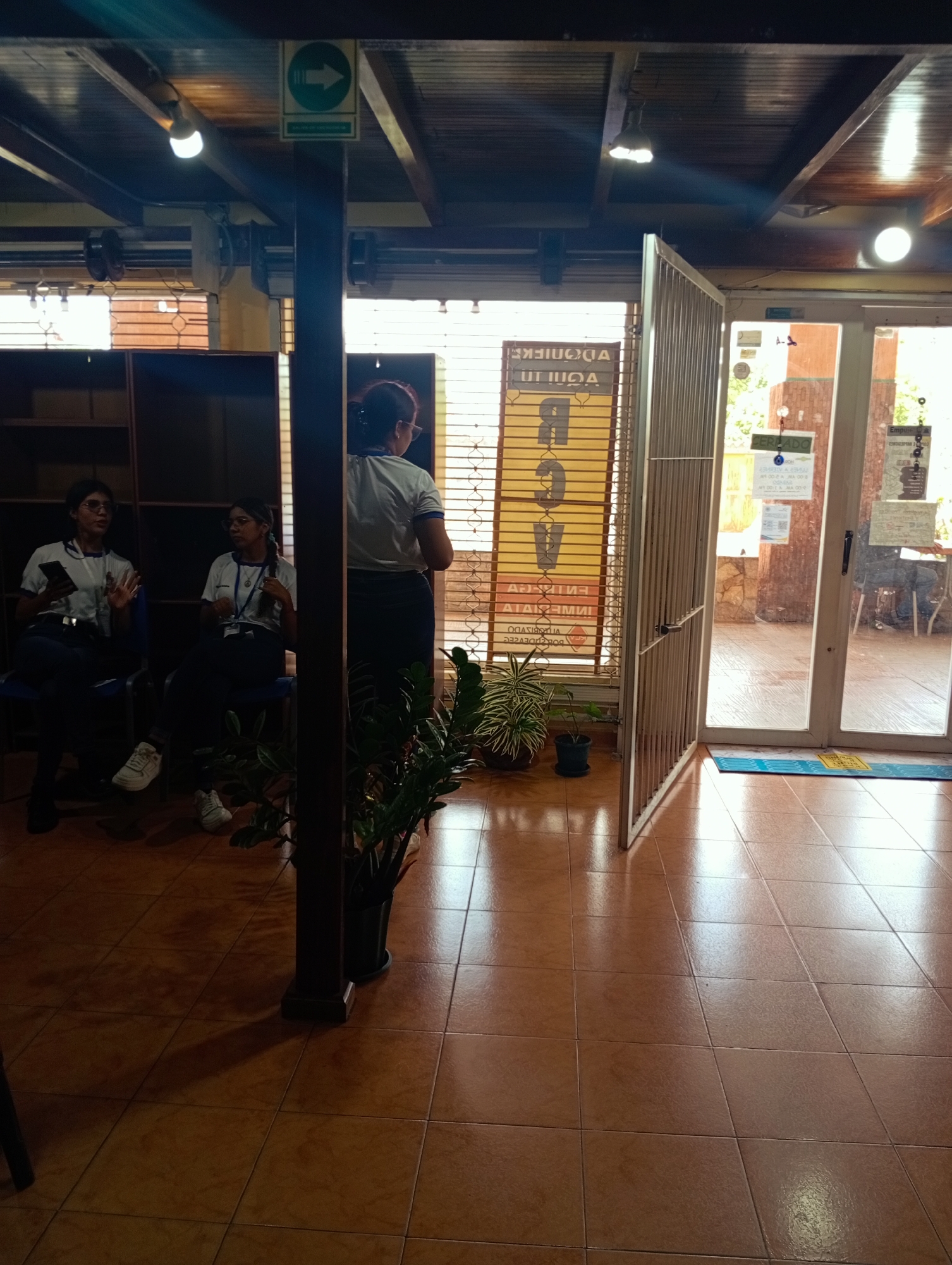 The image depicts an indoor scene, likely a business or office environment, located in Juan Griego, Venezuela. In the foreground, a dark brown tiled floor reflects light. A dark vertical pillar stands prominently, casting a shadow on the floor. To the left, two women are seated on dark chairs, facing away from the camera. Both are wearing white polo shirts with blue trim and dark pants. The woman on the left holds a smartphone in her left hand. The woman on the right wears glasses and has a lanyard around her neck. Between them and slightly behind, a large potted plant with green and white leaves is visible.
In the mid-ground, a large, open metal gate with vertical slats stands on the right side of the image, leading to an exterior area. A yellow sign with large black text in Spanish, partially obscured by the gate, is visible to the left of the gate. Part of the text appears to read "UT IIOA" and "ABOGADOS". Beyond the gate, a glass door reveals an outdoor area with trees and a wooden post. A blue and yellow mat is placed on the floor in front of the glass door.
The background shows wooden ceiling beams and lighting fixtures. A green sign with a white arrow pointing right is mounted on one of the beams. Another sign, partially visible above the yellow sign, appears to have "EMPRESA" written on it. The overall lighting suggests it is daytime.