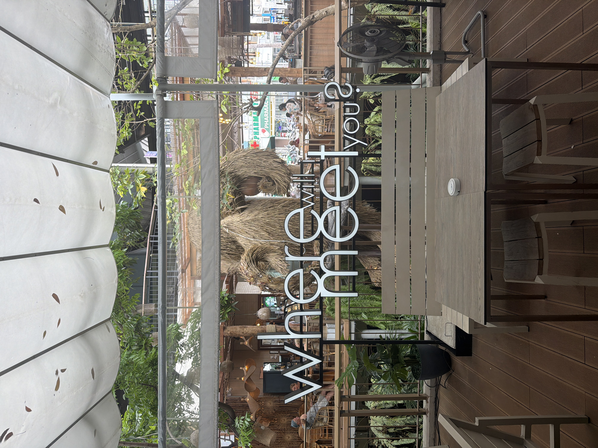 The photo was taken in บ้านบางรักษ์, Thailand. It depicts a patio area with a table and chairs, suggesting a cafe or restaurant setting. The foreground features a sign with the words "Where will we get you?" in a modern, sans-serif font. Through the sign and the surrounding structure, the background reveals a bustling street scene with a pharmacy and various shops, giving a sense of the location being in a commercial area. The patio itself is decorated with plants, including ferns, and there is a large fan providing ventilation. The decking underfoot and the overall design suggest an outdoor seating area with a casual atmosphere.