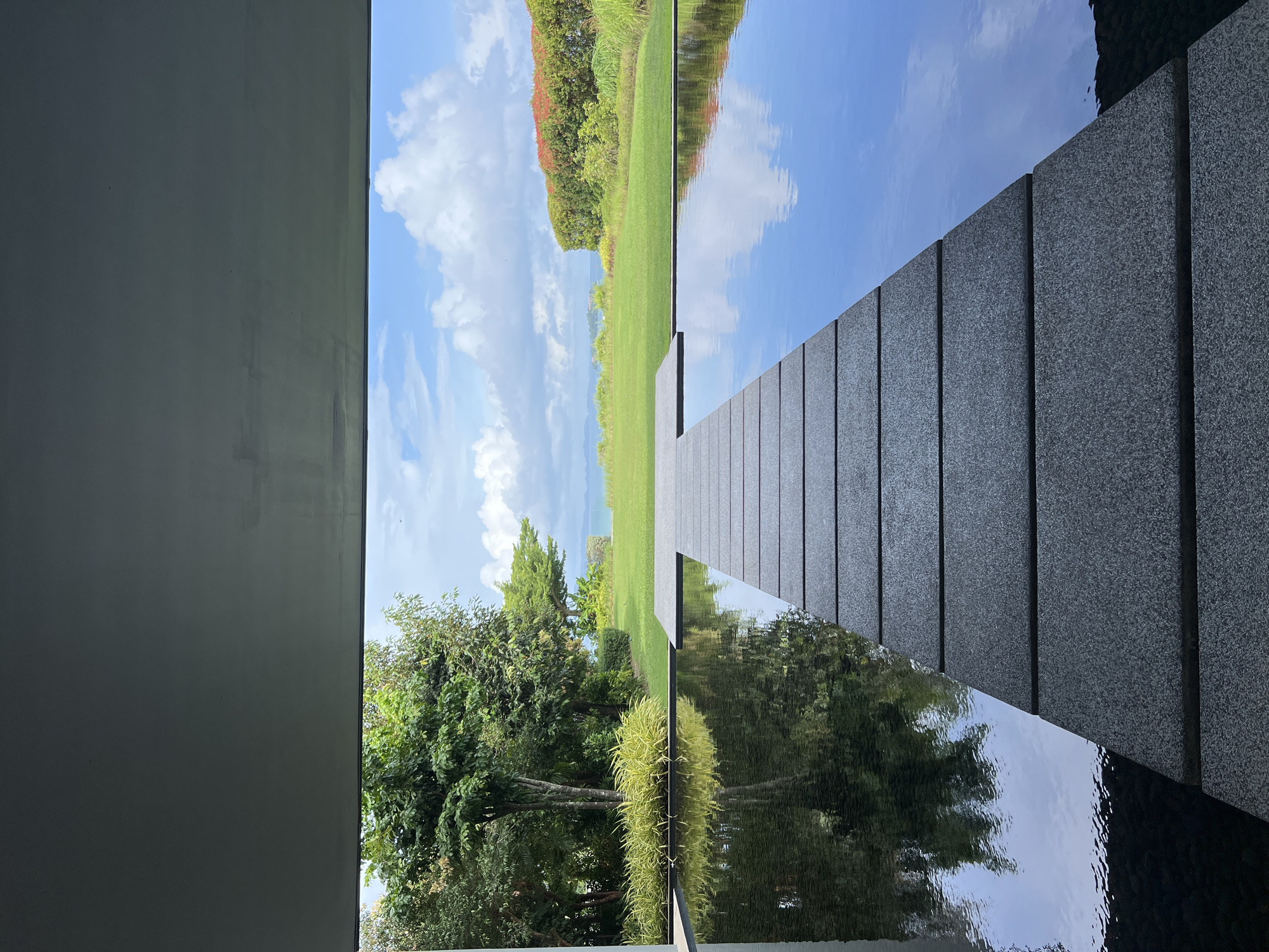 This image depicts a contemporary architectural scene with a focus on water features and landscaping, likely situated in Pa Khlok, Thailand, given the lush tropical vegetation and the distant view of the sea.
In the foreground, a dark, rectangular reflecting pool dominates the lower half of the frame. A pathway of large, grey, rectangular stone slabs is laid across the water, extending from the bottom center towards the horizon. The water is still, creating clear reflections of the surrounding greenery and the sky. The surface of the pool is dark, possibly due to its depth or the material lining it.
Beyond the pool, a manicured green lawn stretches out, leading to a low hedge with red and green foliage. Further in the distance, the sea is visible under a bright blue sky with scattered white clouds. Lush tropical trees with dense green foliage are on the left side of the image, framing the lawn and the distant sea view. The overall impression is one of tranquility and sophisticated design, integrating natural elements with modern architecture. There are no people or animals visible in the image. The scene appears to be taken from within a covered structure, as indicated by the dark ceiling and the framing of the view.