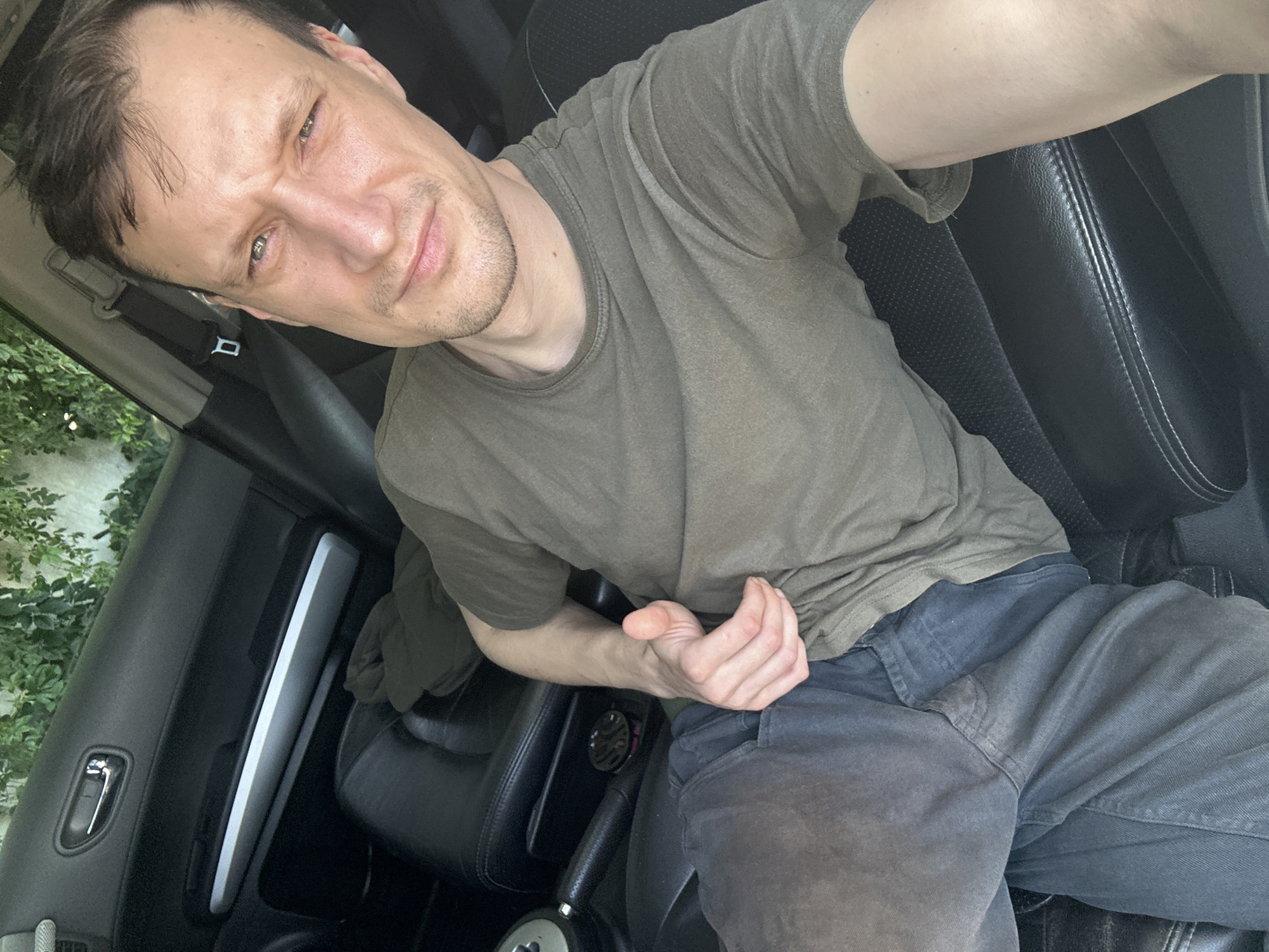 A man is sitting in the driver's seat of a car, possibly in Shatava, Ukraine. He is wearing a green t-shirt and dark pants. His left hand is resting on his leg, and his right arm is raised, with his thumb up. The interior of the car is visible, including the dashboard and seats. There are green plants outside the car window. The lighting suggests it is daytime.