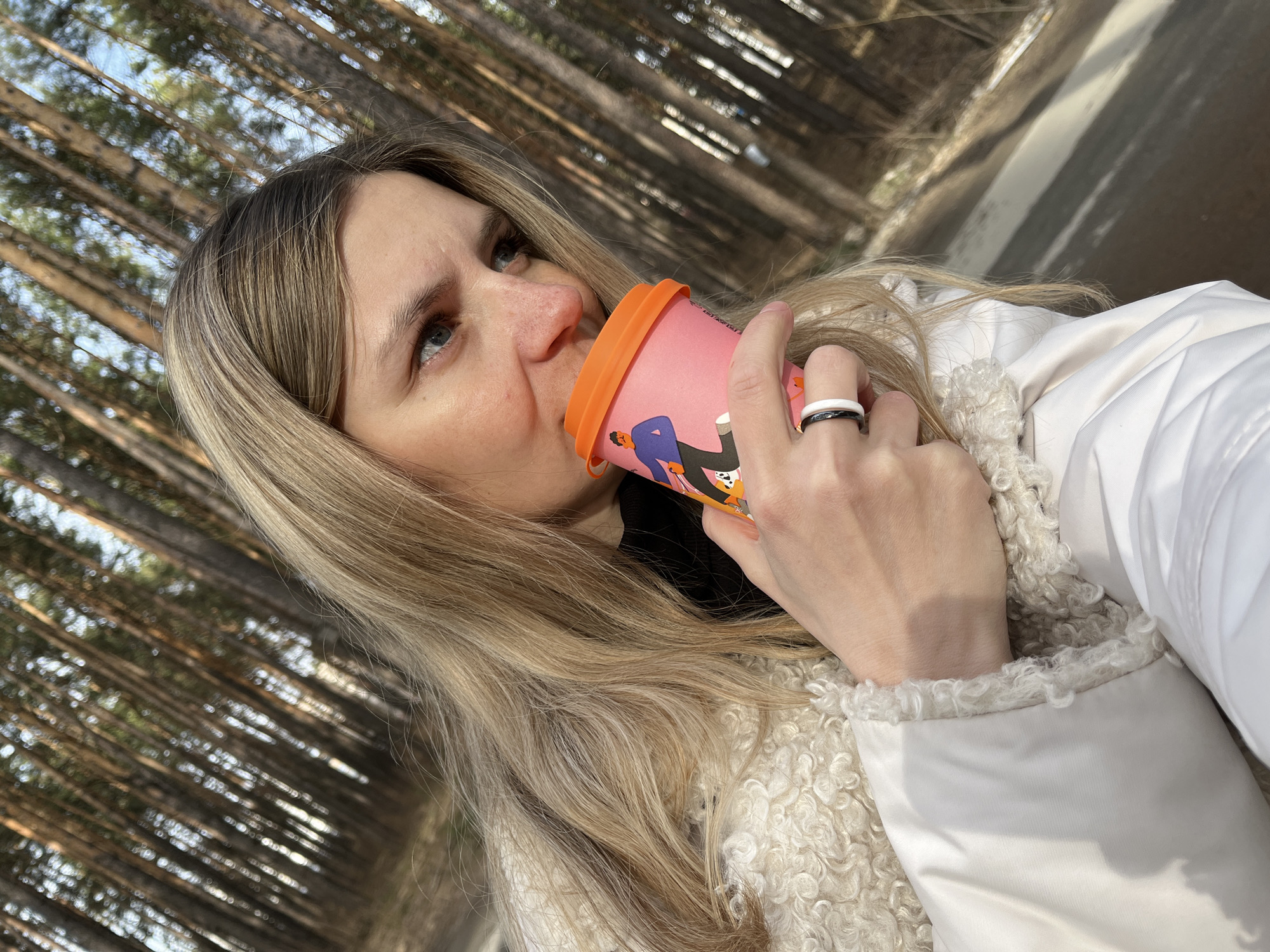 A blonde woman is drinking from a pink and orange patterned cup outdoors. She wears a white coat with a textured, possibly fleece, trim. A black and white ring is visible on her right hand. The background features tall, slender trees, likely in a forest or park setting. The location is Chaikovsky, Russia. A partially visible paved path suggests a walking area. The woman appears to be looking upwards and to the left.