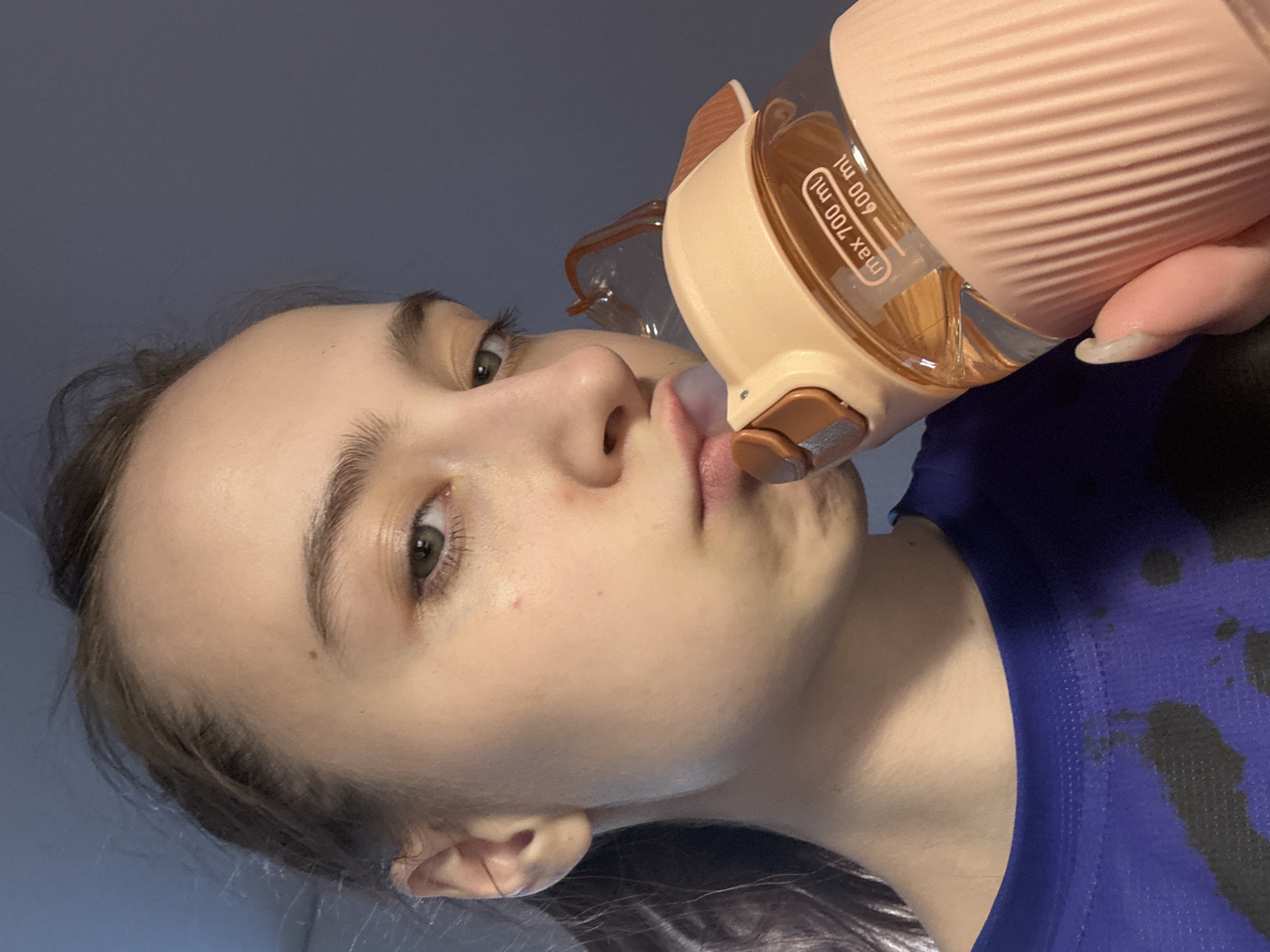 A woman is drinking from a pink water bottle. The bottle has a volume scale labeled "max 700 ml" and "600 ml". She is wearing a blue shirt with dark blotches. The background is a plain, dark color. It is not possible to determine the location in Chaikovsky, Russia from the provided image.
