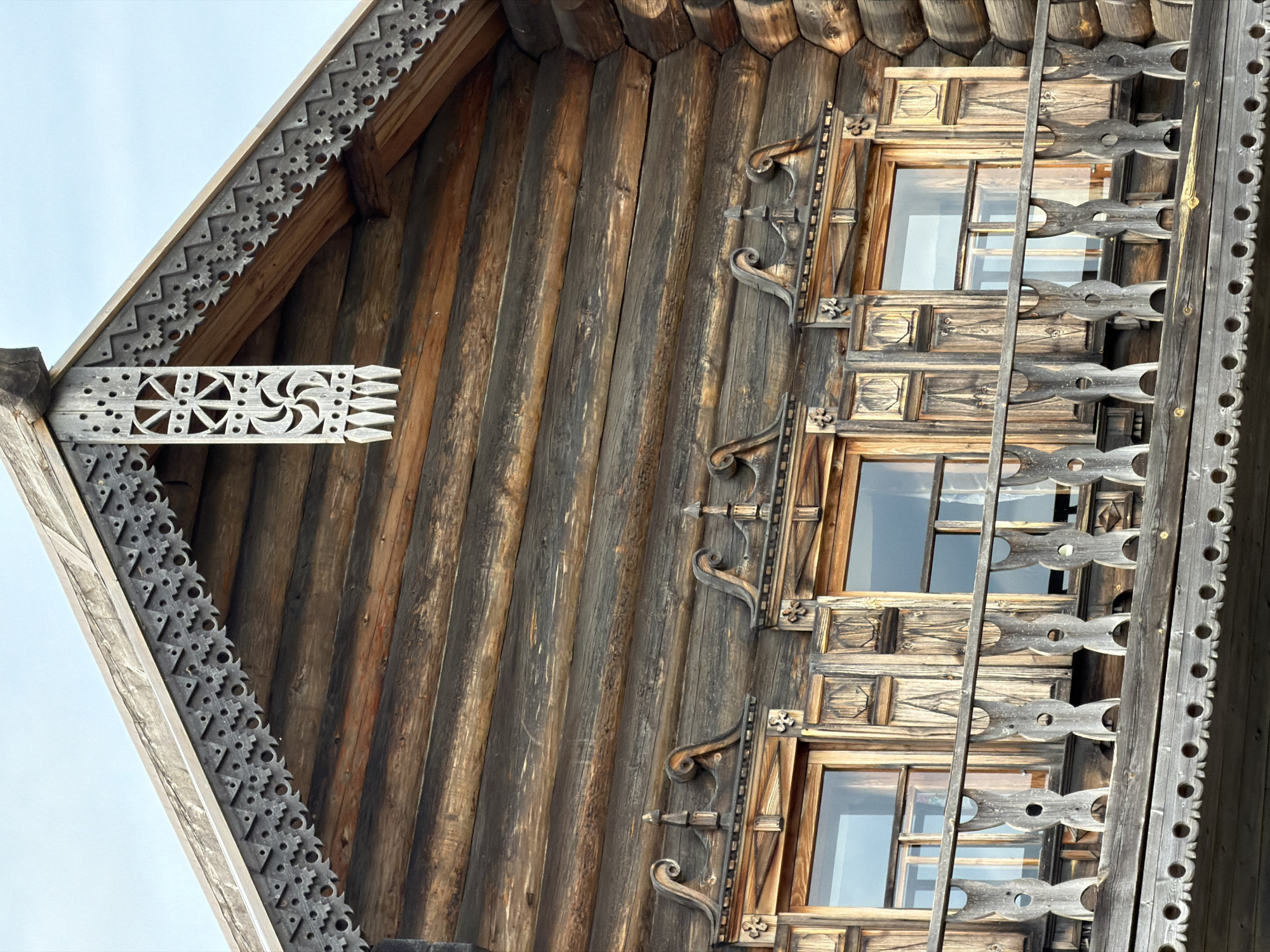 The image displays a close-up view of the upper section of a traditional wooden log house in Dnp Ryzhiki, Russia. The house features intricate, carved wooden trim along the eaves of the roof and around the windows. The walls are constructed from horizontally stacked, rough-hewn logs. There are three windows visible, each with wooden shutters and ornate frames. Below the windows, a wooden balcony railing with carved detailing runs across the facade. The sky is visible in the upper portion of the image. No people are present. The overall impression is one of traditional Russian architecture and craftsmanship.