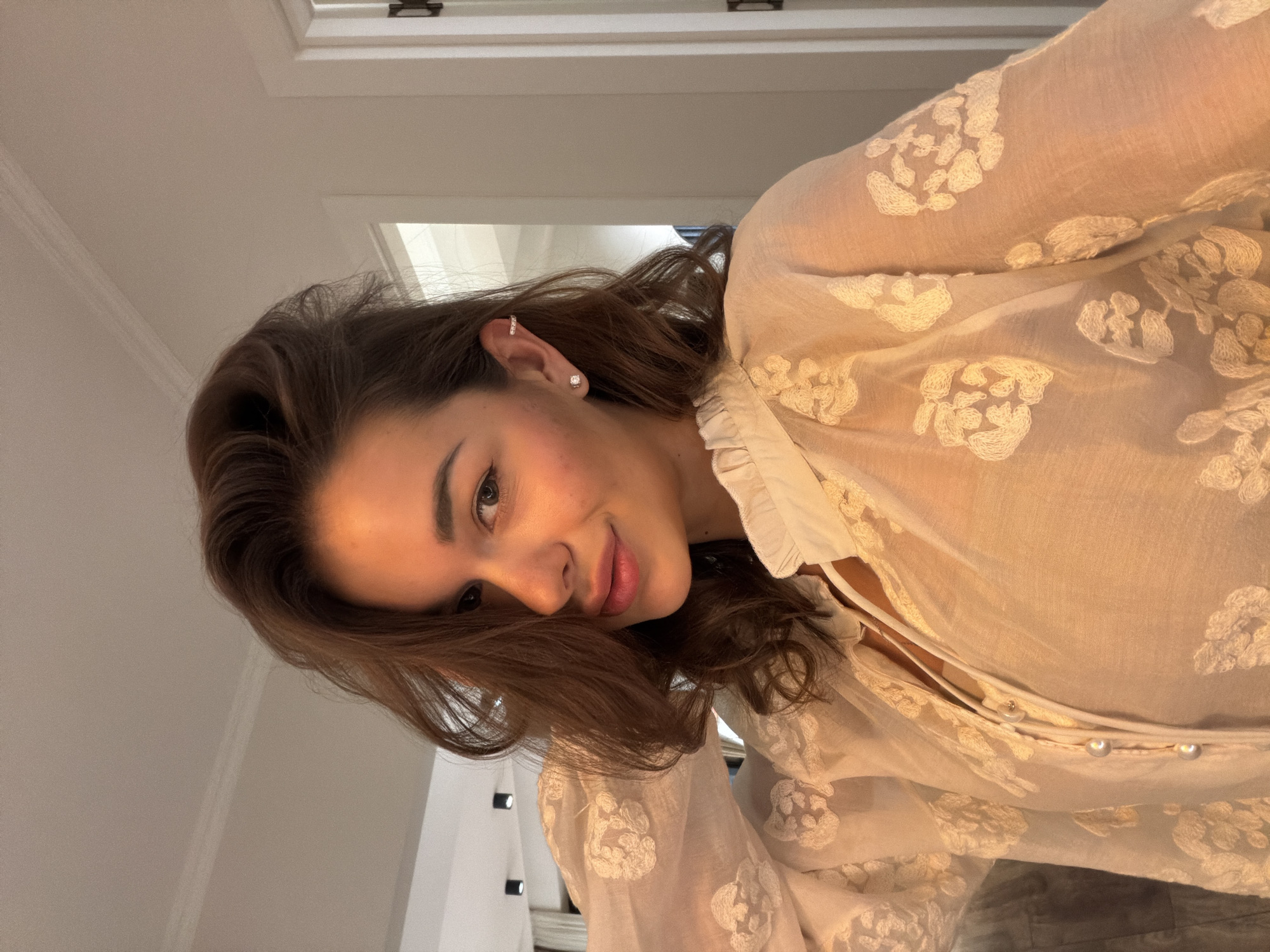 This image captures a close-up, chest-up indoor portrait of an adult female in a residential setting in Tbilisi, Georgia. The subject has long, wavy, brown hair, with some strands partially covering the right side of her face (viewer's left). She is looking directly at the camera with a slight smile. Her visible right ear (viewer's left) is adorned with a stud earring in the lobe and a small cuff earring on the upper cartilage. She is wearing a light-colored, possibly cream or white, sheer blouse featuring a high ruffled collar and intricate white floral embroidery. Three pearl buttons are visible on the central placket of her blouse. Her left arm (viewer's right) is raised, with her hand positioned near her head, suggesting a posed action. The background features light-colored walls and a ceiling with white crown molding. A white paneled door with a dark handle or hinge is visible to the right. In the distant left background, modern dark spotlights are present on the ceiling of an adjacent interior space. The scene is illuminated by warm, artificial light.