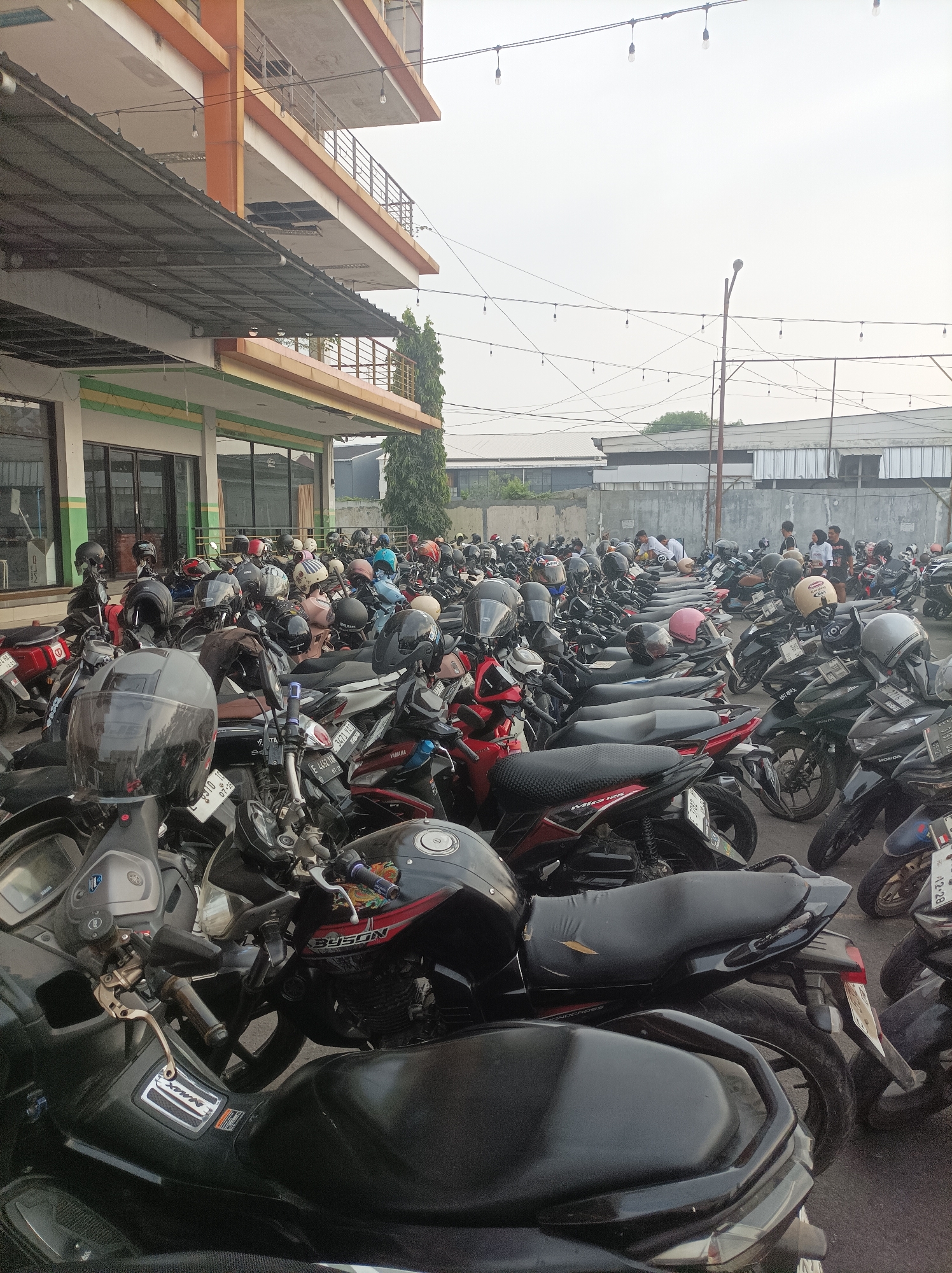 The image depicts a large outdoor parking lot filled with numerous motorcycles, many with helmets placed on them. The parking lot appears to be adjacent to a multi-story building with an orange and white facade, featuring balconies and metal railings. String lights with exposed bulbs are visible overhead, crisscrossing the sky, which is overcast and hazy. In the background, several individuals are standing and interacting, and more buildings and structures can be seen. The scene suggests a busy public area in the city of Cirebon, Indonesia, likely a commercial or public facility with a high volume of motorcycle traffic. The overall impression is one of a functional, everyday scene in an urban Indonesian environment.