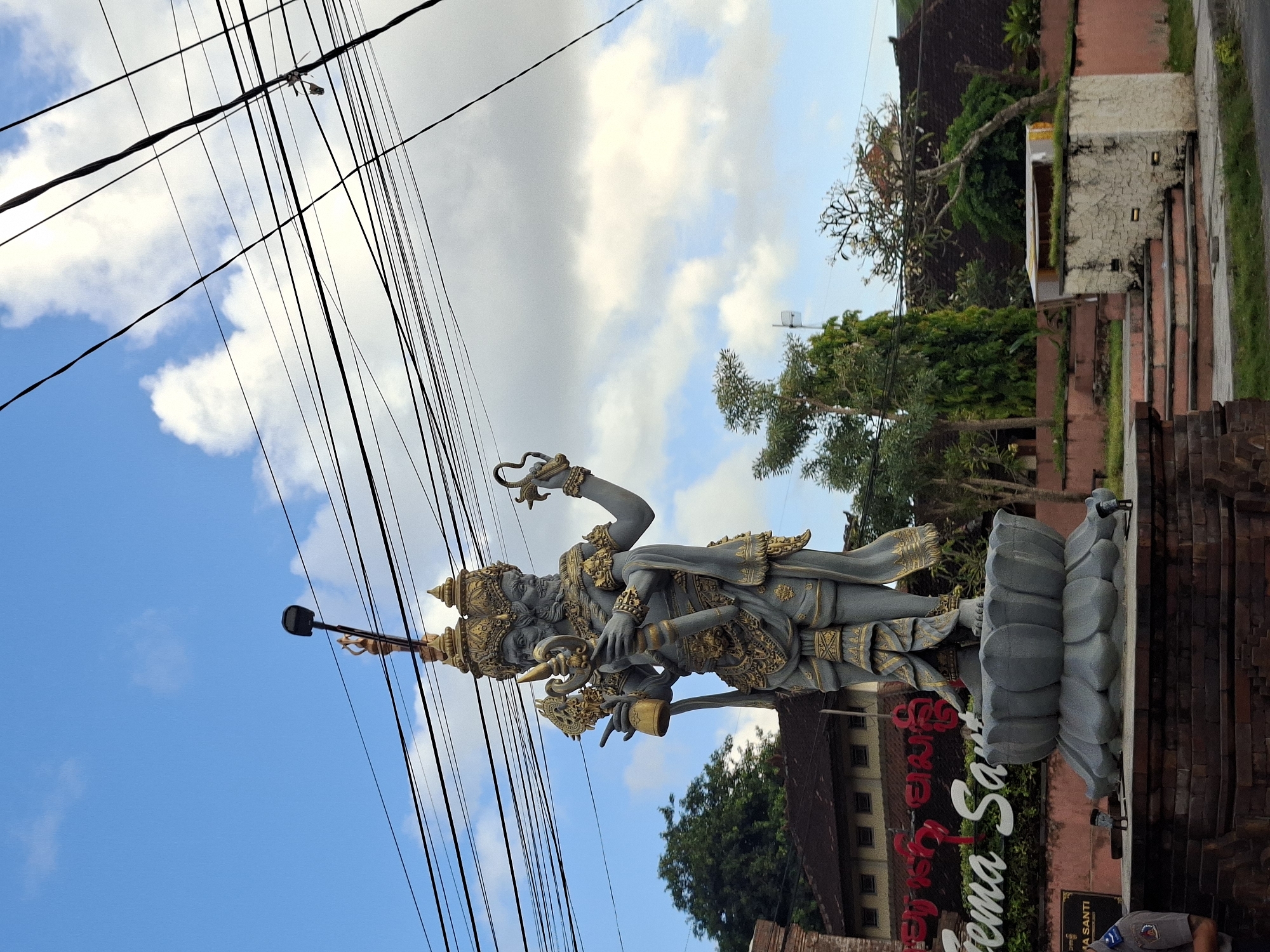 In Nusa Penida, Indonesia, a large stone statue depicting a multi-headed deity stands prominently on a lotus-shaped pedestal. The statue is intricately adorned with gold accents and holds various symbolic objects. Behind the statue, a building with a red roof and a sign displaying text in a non-English script is partially visible. Overhead, a complex network of power lines crisscrosses the sky, which is filled with scattered white clouds against a blue backdrop. The foreground shows a brick base supporting the pedestal, with some grass and paving stones around it. To the left, a portion of a brick wall and a sign with the text "SANTI" is visible. To the right, there are stairs leading up to a structure with a white facade.