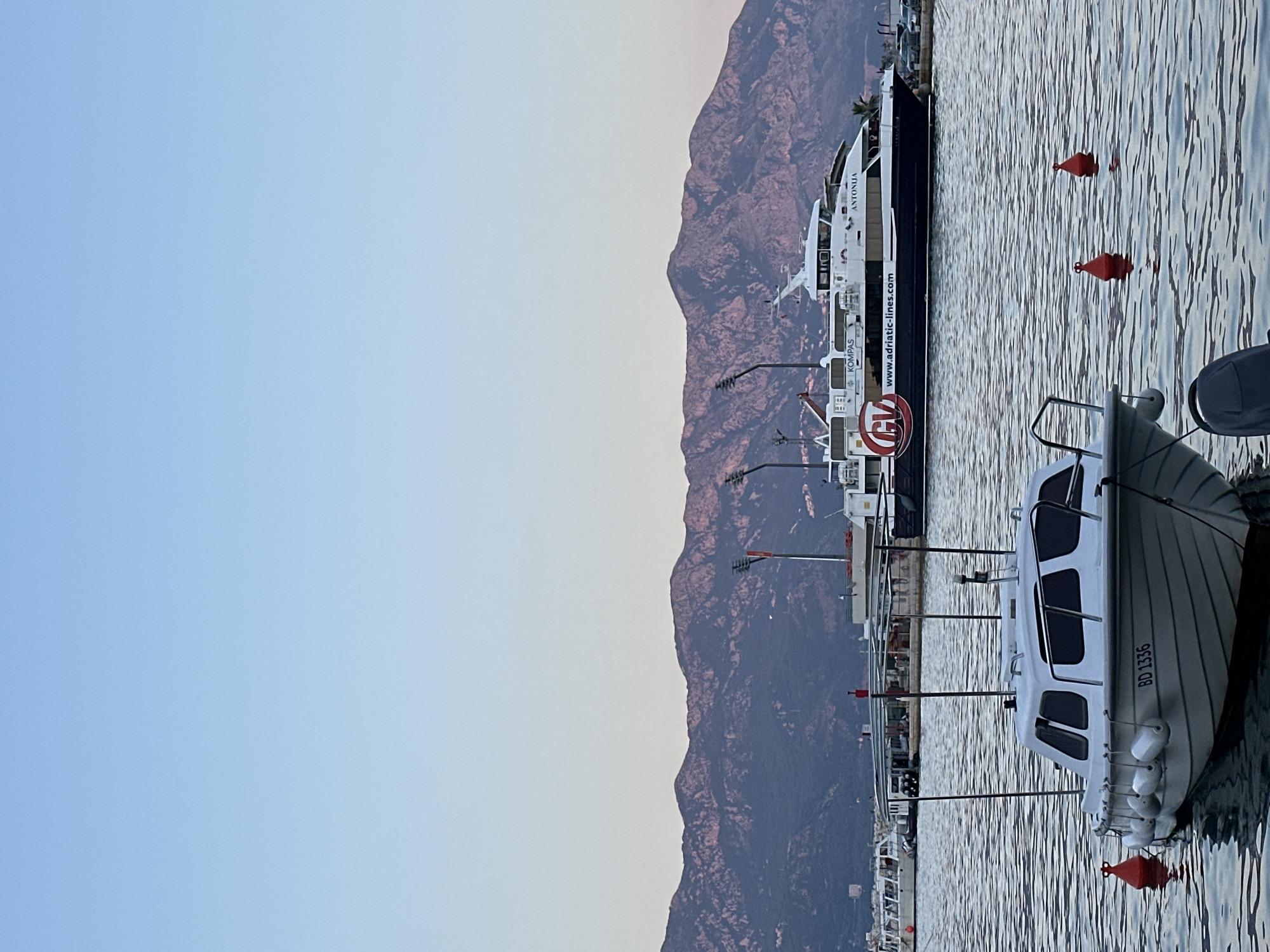 The image depicts a serene harbor scene in Budva, Montenegro, with a clear blue sky and a range of mountains in the background. In the foreground, a small white boat with the registration number "BD 1336" is moored. The boat has a white cabin with tinted windows and is surrounded by several red buoys.
Further in the harbor, a larger ferry named "ANTONIJA" is docked. The ferry is white with a dark blue hull and features the website "www.adriatic-lines.com" and the logo "GV" printed on its side. Other vessels, including what appears to be a passenger boat, are also visible in the background. The water is calm, with gentle ripples reflecting the sky and the surrounding objects. The mountains in the distance are dusky, suggesting either early morning or late evening light. The overall impression is one of tranquility and a typical day in a coastal town.