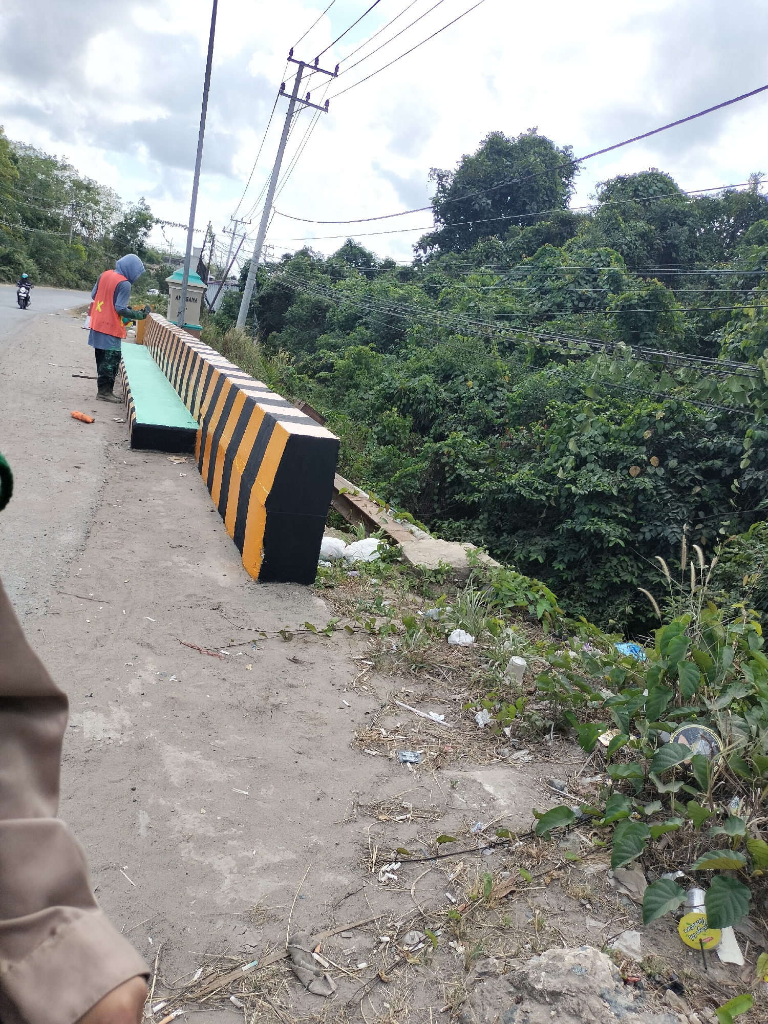 In Angsana, Indonesia, a person wearing an orange safety vest and a blue hood is painting a concrete barrier along a road. The barrier is painted with alternating black and yellow stripes, a common safety marking. The person appears to be using a roller to apply the paint, with a small orange object, possibly a paint can or roller handle, on the ground nearby. To the left of the barrier, a green section has been painted, possibly a bench or a lower part of the barrier. The road appears to be paved with asphalt or concrete. In the background, lush green trees and vegetation cover a hillside, and utility poles with power lines are visible. A motorcycle is parked on the roadside in the distance. The ground beside the barrier is unpaved and appears to have some debris, including white plastic bags and dried vegetation. The sky is overcast with clouds.