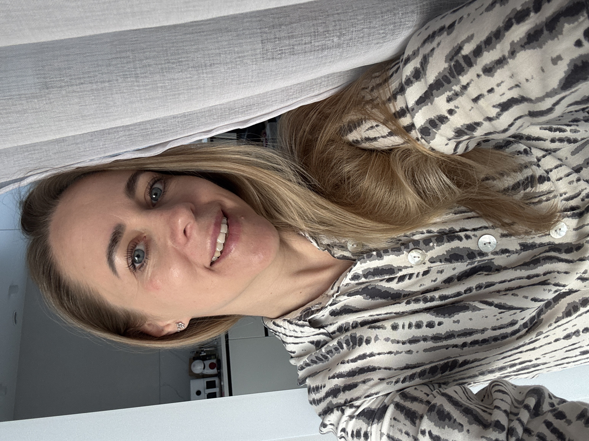 A woman with blonde hair and blue eyes is smiling at the camera. She is wearing a patterned shirt with buttons. The background includes a light-colored curtain and a glimpse of a kitchen with appliances and cabinets. The location in the city of Chaikovsky, Russia, is not discernible from the image.