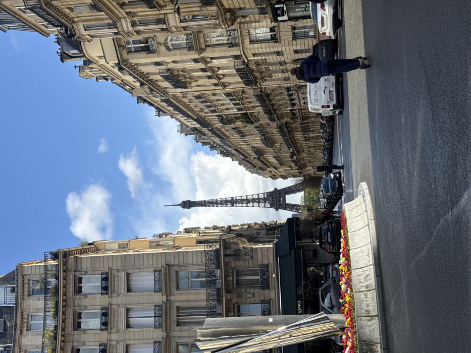 The image displays a wide Parisian street leading toward the Eiffel Tower in Paris, France, under a partially cloudy blue sky.
In the foreground, to the left, a curved, raised concrete planter filled with colorful flowers (red, yellow, orange, pink) occupies part of a central island. Behind this, a tall, multi-faceted metallic sculpture stands. Adjacent to the sculpture is a building with a dark green awning displaying "VINO," part of a larger Haussmannian structure. A silver car is parked near this area.
The street, paved with asphalt, extends into the midground. On the right side, a person wearing a dark blue hooded jacket is riding an electric scooter away from the viewer. Further along the right curb, a white commercial van marked "idex" is parked, followed by a line of other parked cars. On the left side of the street, another person in dark clothing stands near parked vehicles, including a dark SUV, with several indistinct figures visible further down.
The background is dominated by the Eiffel Tower, centrally positioned and framed by rows of classic Haussmannian buildings on both the left and right sides of the street. These buildings feature multiple stories, light-colored stone facades, numerous windows with dark wrought-iron railings, and mansard roofs. Some upper-story balconies exhibit greenery. The base of the Eiffel Tower is partially obscured by trees. Shadows are cast across the street, indicating daylight conditions.