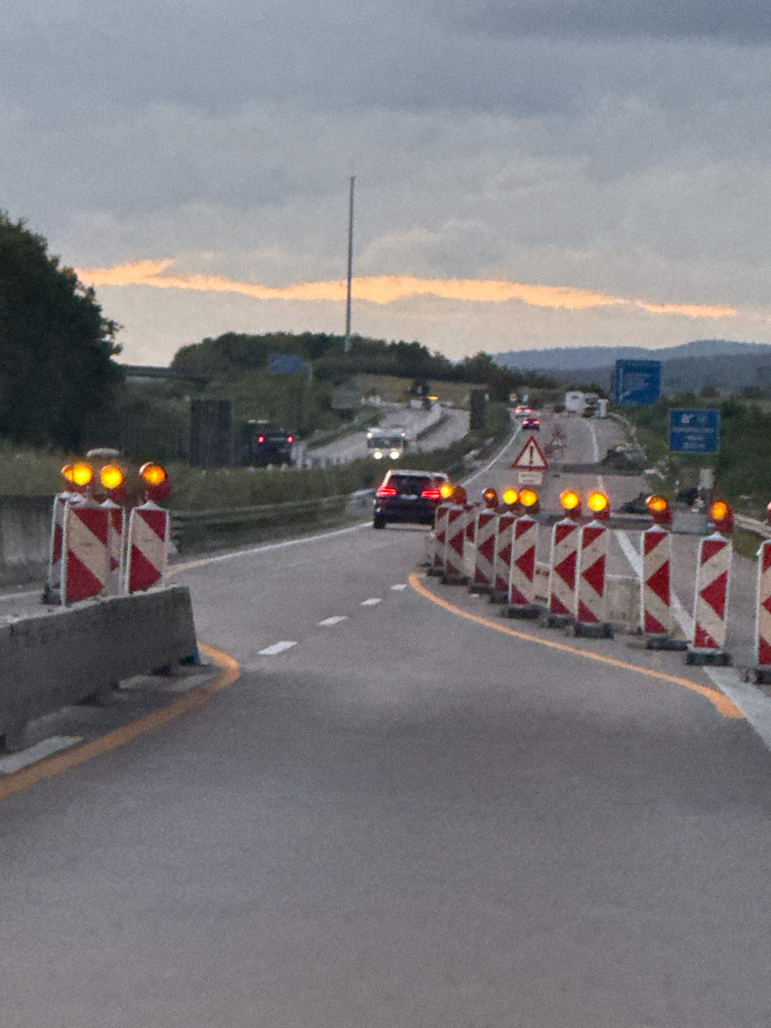 The image depicts a highway scene in Werther, Germany, during dusk. A line of construction barriers, marked with alternating red and white diagonal stripes and topped with flashing orange lights, guides traffic around a closed lane. The road surface is asphalt, with a yellow line delineating the edge of the active lane and white dashed lines marking the lane divisions. Several vehicles are visible on the highway, including a dark-colored car in the foreground that appears to be driving in the active lane, and other cars and trucks further in the distance. The sky is overcast with a strip of orange light visible on the horizon, suggesting the sunset. The background consists of green trees and hills, with a tall tower or antenna visible on the right side of the frame. The overall impression is one of a construction zone on a major roadway under low-light conditions.