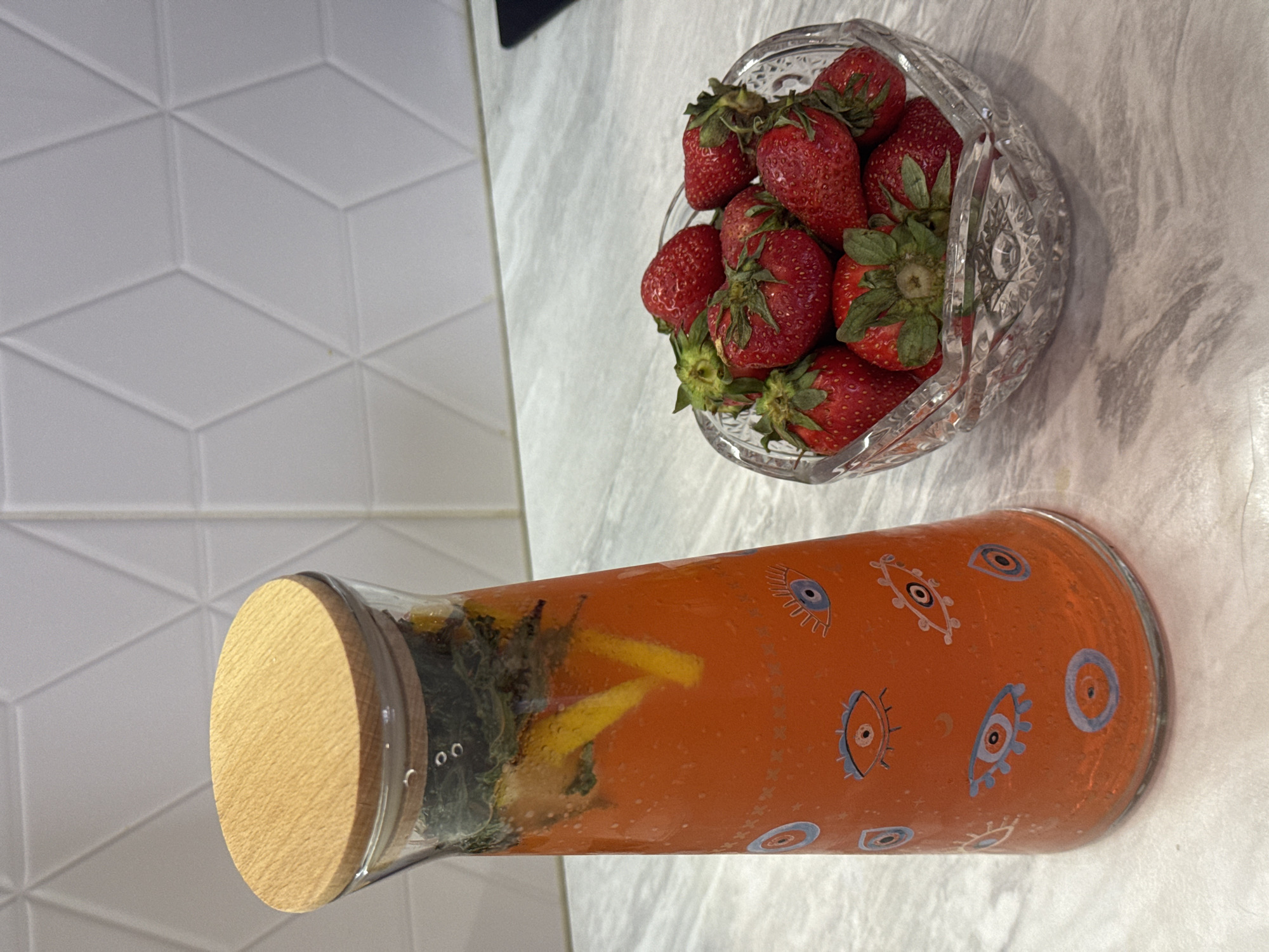 The scene depicts a counter, possibly in a kitchen or dining area. A glass bowl filled with strawberries sits on the right side of the frame. A glass pitcher with a wooden lid, containing a liquid and decorated with eye-like symbols, lies horizontally beneath the bowl. The background includes a wall with a geometric tile pattern. There's no evidence to suggest this scene is located in Izhevsk, Russia. No people are present in the image.