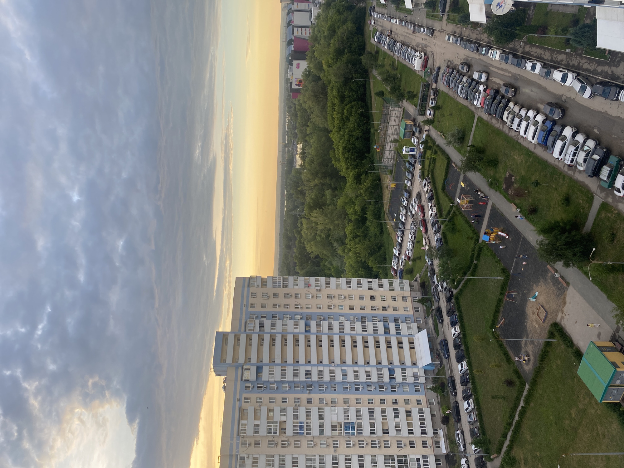 The image depicts an aerial view of a residential area in Novokuybyshevsk, Russia, during a sunset. A large, multi-story apartment building with light yellow and blue facade dominates the left side of the frame. In front of the building, a parking lot is filled with numerous cars. To the right of the apartment building, a dense forest separates the residential area from a distant cityscape that includes several commercial buildings. A playground with swings and a sandbox is visible in the foreground, with a few people present. A small utility building with a green roof is situated in the lower left corner. The sky is partly cloudy, with soft hues of yellow and orange indicating the setting sun.