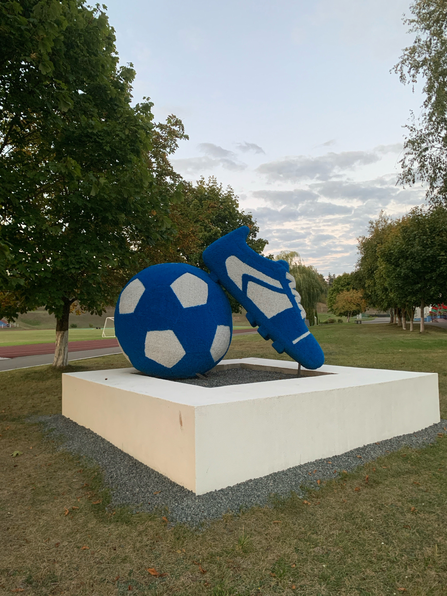 The image shows an outdoor sculpture in Kapyĺ, Belarus, depicting a large blue soccer ball and a blue soccer cleat. The sculpture is placed on a white, rectangular pedestal that is surrounded by gravel. The background features green trees, a grassy field, and a running track, suggesting the sculpture is located in a park or near a sports facility. The sky is overcast with some clouds. There are no people visible in the image.