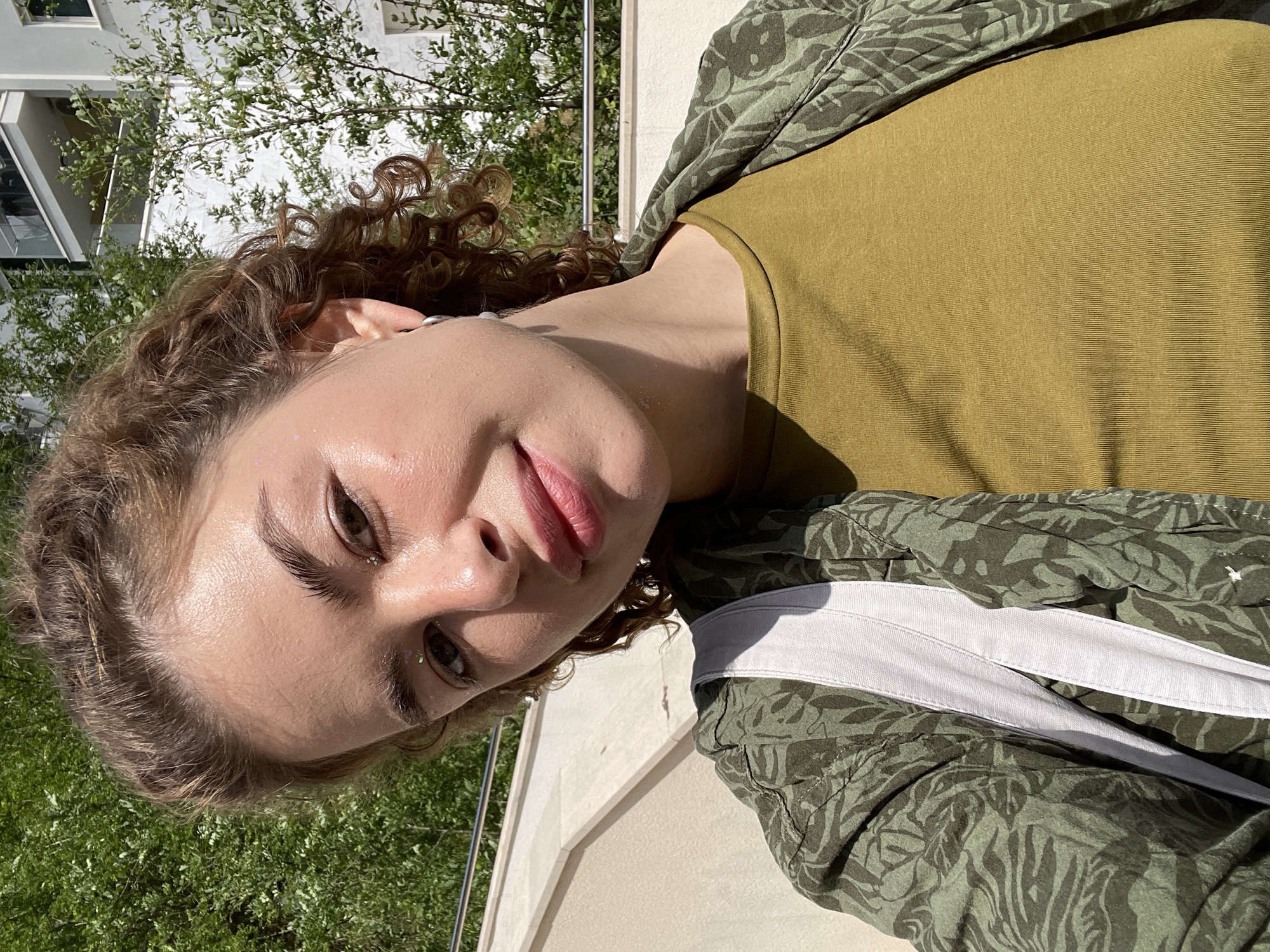 A portrait of a woman with curly hair, wearing a green shirt and green patterned jacket with a white stripe. She is looking at the camera with a slight smile. Her skin is dewy, and she has glitter on her face. The background shows green foliage and a building with white walls, indicating an outdoor location in a developed area. The location of Boreti, Montenegro, is not directly discernible from the image.