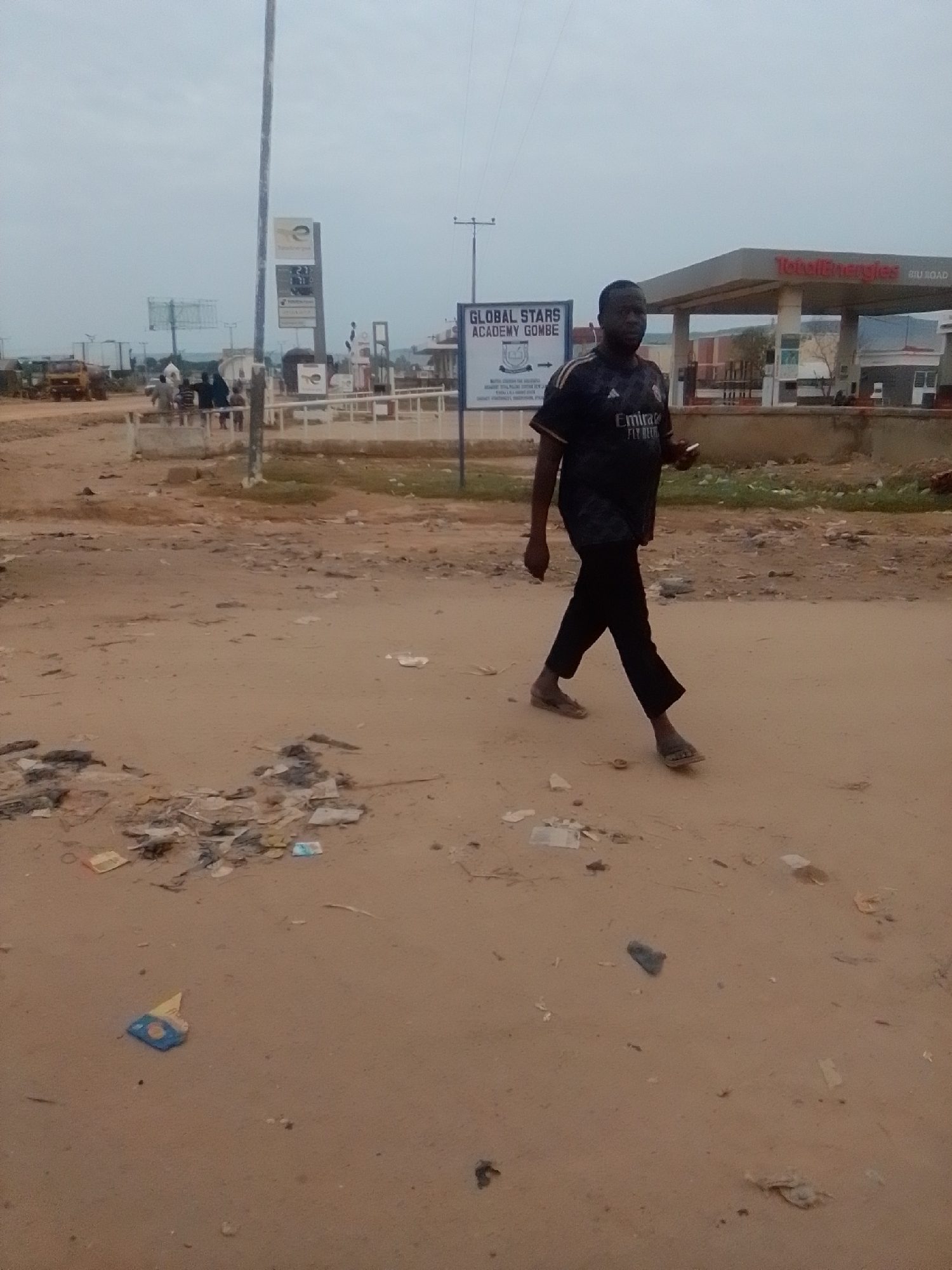 The image depicts a dusty, outdoor scene in Gombe, Nigeria. In the foreground, a man walks from right to left across a dirt road littered with debris, including paper and plastic waste. He is wearing a dark t-shirt with "Emirates Fly Better" written on it, black pants, and sandals. He holds a mobile phone in his right hand.
In the background, a TotalEnergies gas station is visible, with the company's red and white branding prominently displayed. A sign for "GLOBAL STARS ACADEMY GOMBE" is also present, with text that appears to be academic information and a crest. Other elements of the background include a tall pole, some trucks, and a few indistinct figures standing near a low wall. The sky is overcast. The overall environment suggests an urban or semi-urban area with ongoing development or maintenance challenges, indicated by the accumulation of trash on the ground.