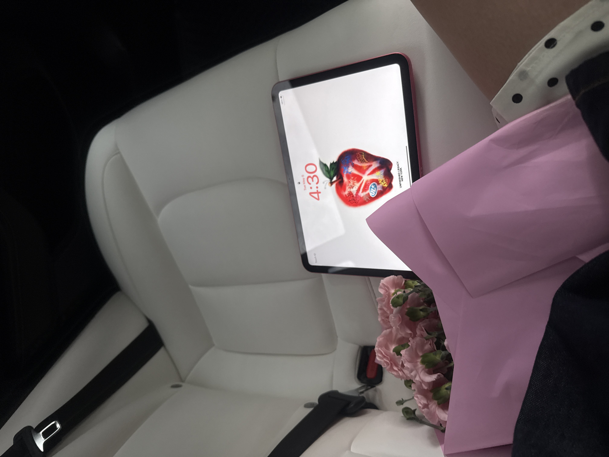A red iPad is displaying a screen showing the time 4:30, with a decorated apple icon, resting on a white car seat next to a bouquet of pink carnations wrapped in pink paper. The car seat has a black seatbelt, and the interior of the car appears clean and modern. A person wearing a white collared shirt with black polka dots is partially visible next to the flowers. The image does not provide enough context to determine the location as Yerevan, Armenia.