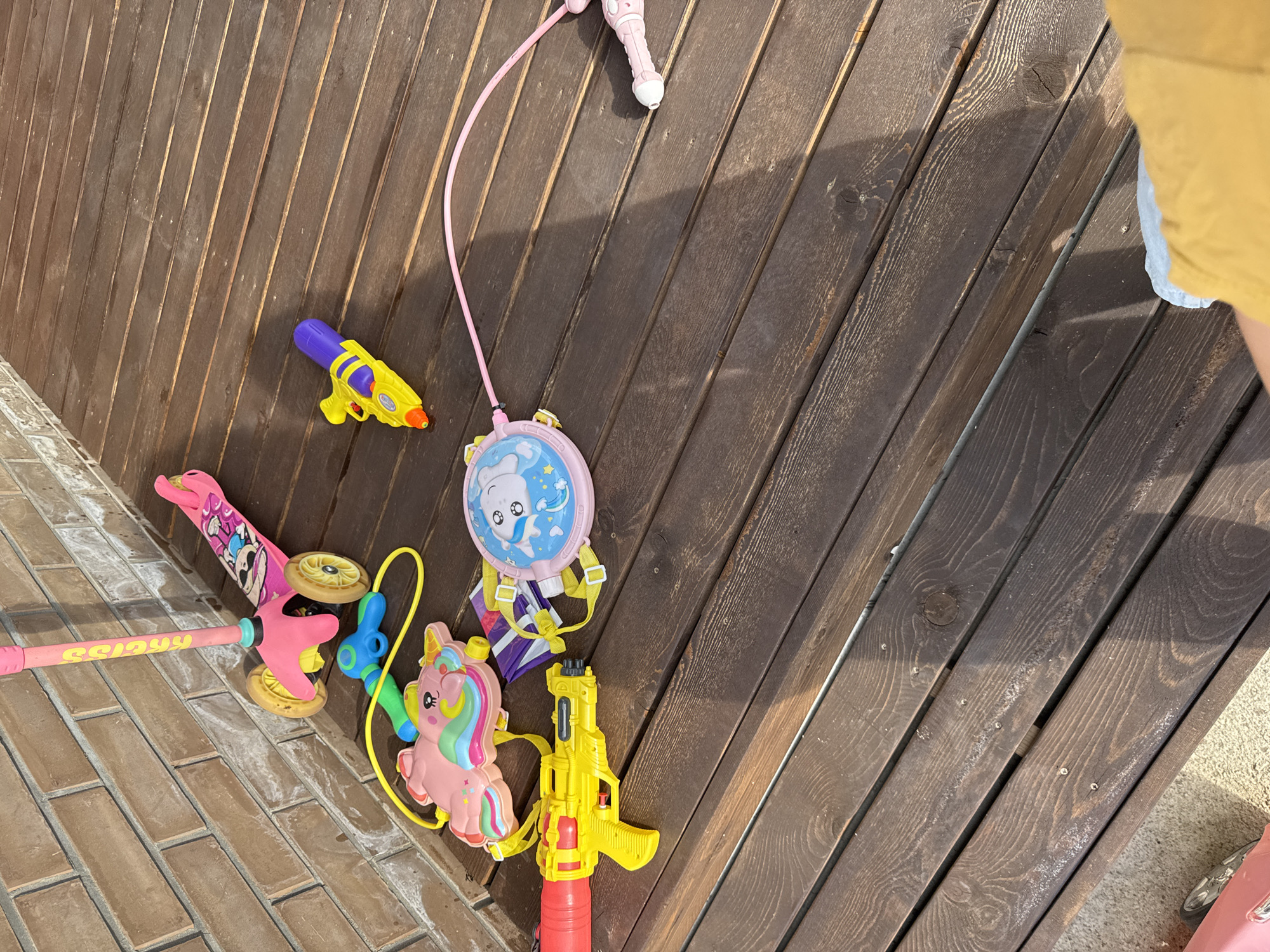 The image shows a collection of children's toys scattered on a wooden deck outside. In the upper left, a pink three-wheeled scooter with a cartoon character design leans against a brick wall. To the right of the scooter, a yellow and purple water gun lies on the deck. Below the scooter, a pink unicorn-shaped water backpack with a yellow strap is visible. Next to the unicorn backpack is a pink circular water shield with a cartoon character. A large yellow water gun with a red reservoir is in the lower left of the frame, positioned on the deck. A pink water gun with a white tip extends from the right side of the image, connected by a pink tube to the water shield. The wooden deck appears weathered and is bathed in sunlight, casting shadows. The background consists of a brick wall and a hint of a gravel or concrete surface on the far left. The scene is set outdoors, likely in a residential area or a park.