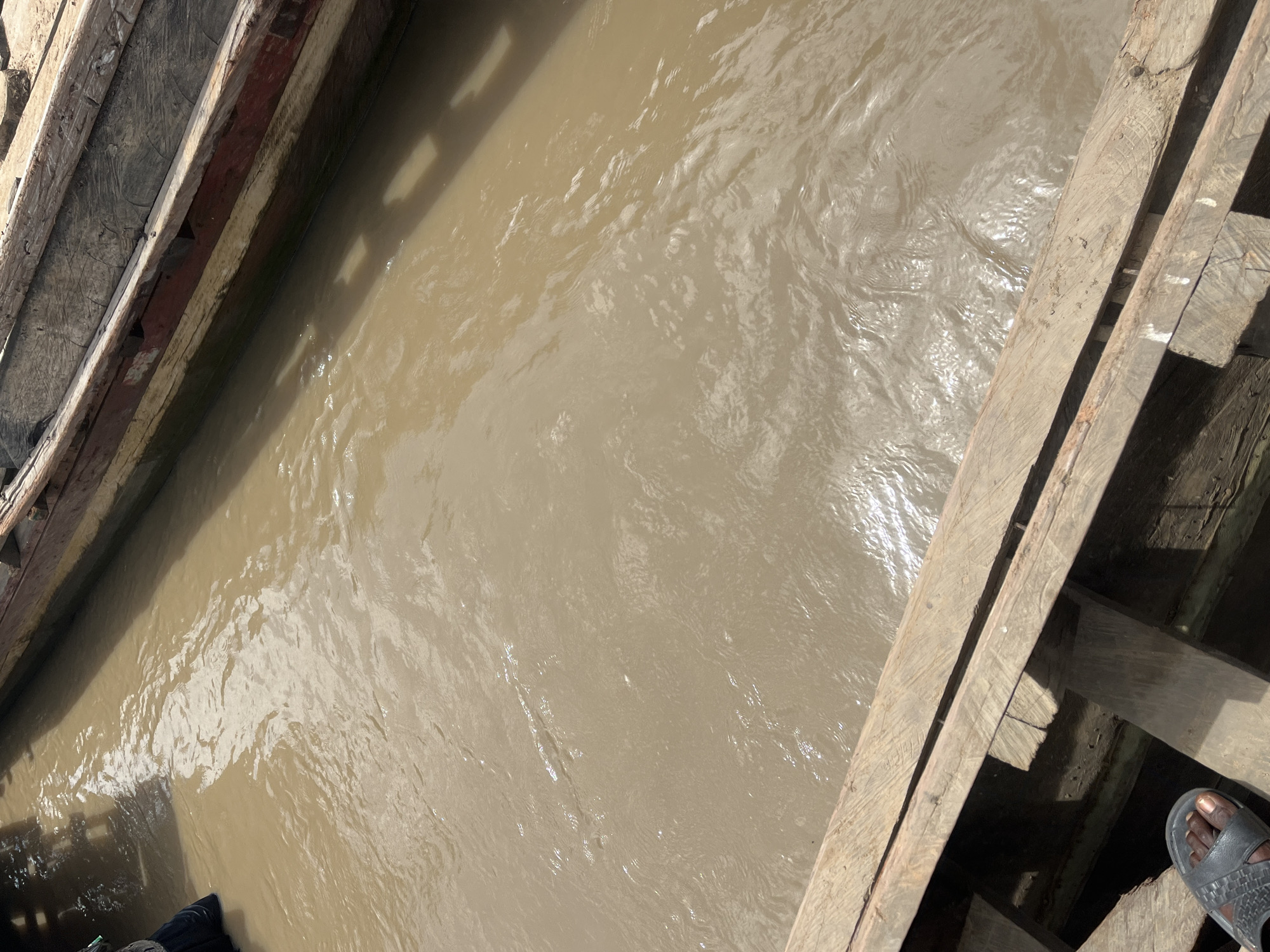 An aerial shot shows a section of the River Benue in Nemnei, Nigeria, with two wooden boats docked alongside each other. The water is a muddy brown, with ripples and sunlight reflecting off its surface. The boats appear weathered, with visible planks and aged wood. In the foreground, a person's foot wearing a black sandal is resting on the edge of one of the boats. A person wearing dark clothing is partially visible in the upper left corner of the image, seemingly standing in or near the water. The scene suggests a typical day on the river, with boats and human activity.