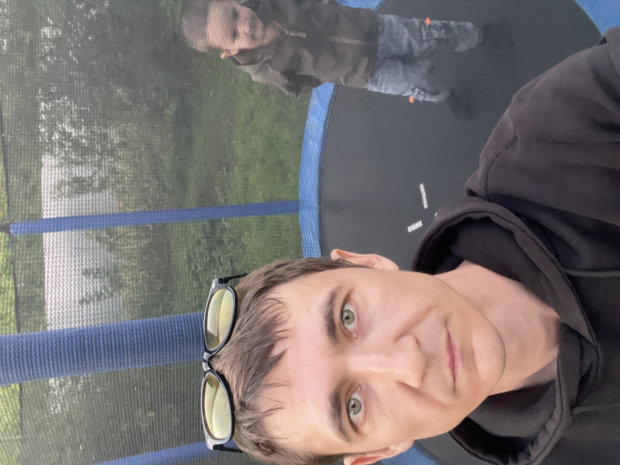 The image shows two people on a trampoline outdoors. The person in the foreground, who appears to be an adult male, is taking a selfie. He has short brown hair, green eyes, and is wearing a black hoodie and sunglasses perched on his head. In the background, a young child, possibly a boy, is visible on the trampoline. He is wearing a dark colored hooded jacket and light colored pants. The trampoline has blue padding around the edge and a black jumping mat. A safety net surrounds the trampoline. The background is a blurred green area, suggesting a grassy yard with some trees or bushes. The setting appears to be a residential area in Laishevo, Russia.