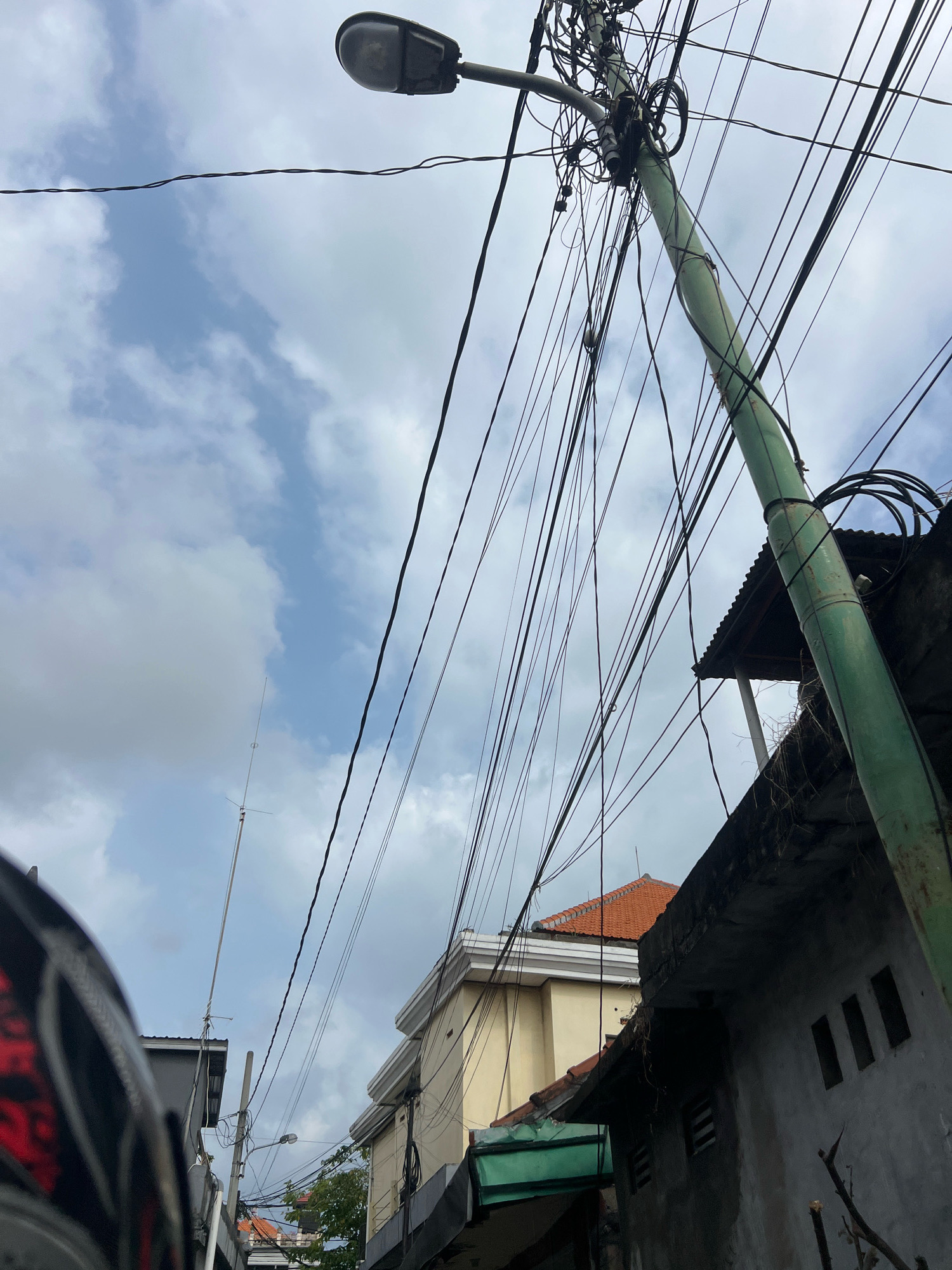 This is an outdoor, eye-level, medium shot photograph taken in Kuta, Indonesia, on a cloudy day. The composition is dominated by a large number of tangled electrical wires crisscrossing against a pale blue sky with scattered white clouds. A tall, green utility pole, heavily laden with wires, stands prominently on the right side of the frame, leaning slightly. A street lamp is attached to the top of this pole. In the background, the rooftops of several buildings are visible, including a building with an orange-tiled roof. The lower left corner of the image shows a portion of a dark-colored motorcycle helmet with a red and black patterned design. The overall impression is of a dense urban environment with an overabundance of overhead electrical infrastructure. There are no people visible in the frame.