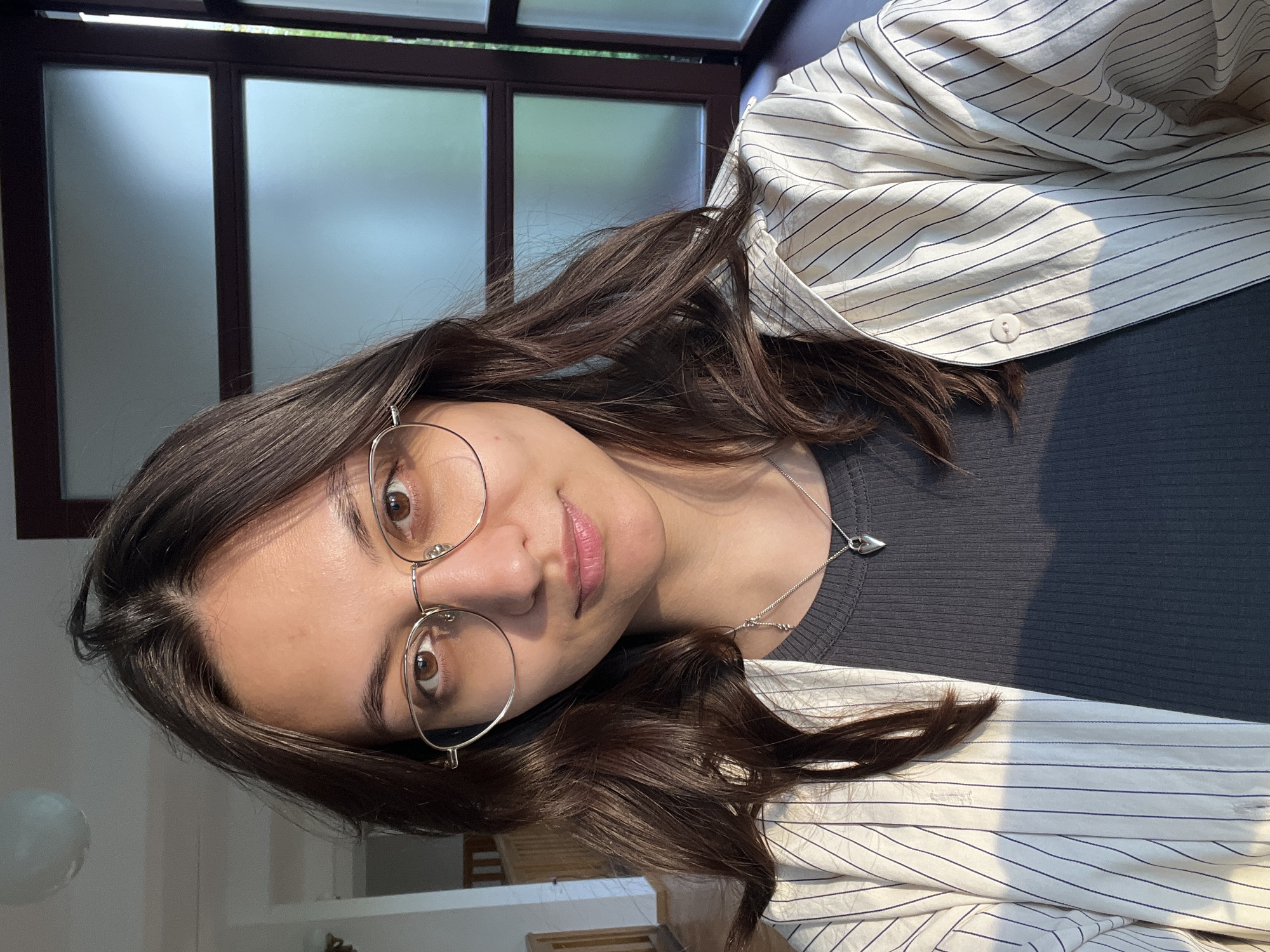 A young woman is posing for a portrait indoors. She has dark brown hair, wears round glasses, a gray ribbed top, and a white and blue striped blazer. She is also wearing a silver necklace with a heart-shaped pendant. Sunlight is streaming in through a window with a dark frame. A glimpse of a room interior is visible in the background, including a light fixture and some furniture. The location cannot be determined to be Yekaterinburg, Russia, based on the image.
