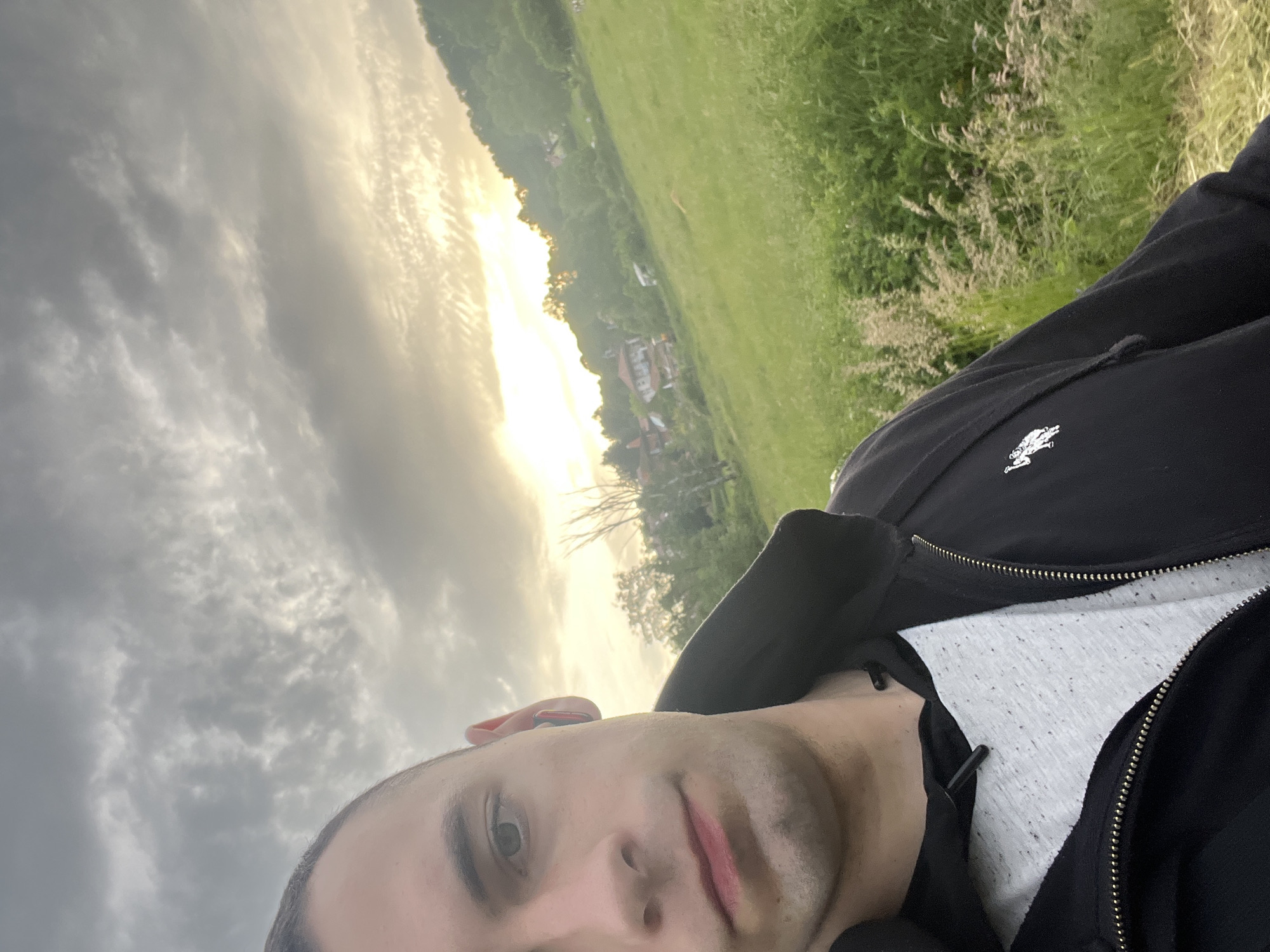 The image shows a selfie of a person with light skin, close-cropped hair, and wearing a black jacket over a light-colored shirt. The background features a landscape with a field, trees, and houses in the distance. The sky is filled with clouds, and the sun appears to be setting or rising, casting a bright light. The location and city are unknown.