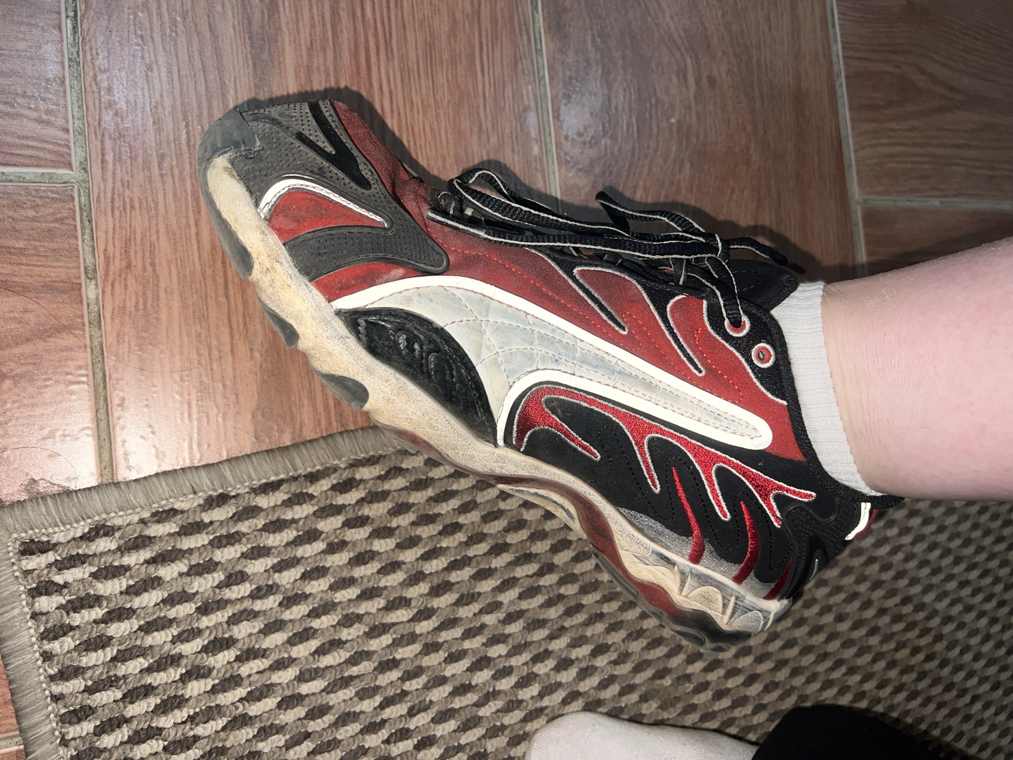 A close-up shot shows a person wearing a well-worn athletic shoe with a predominantly red, black, and white color scheme. The shoe features decorative stitching and an intricate design pattern. The sole is heavily soiled, suggesting frequent use. The person is wearing a white sock. The shoe is situated on a section of patterned carpet and also visible is the reddish tile floor behind the carpet.
There is no information indicating a location in Noviy, Russia.