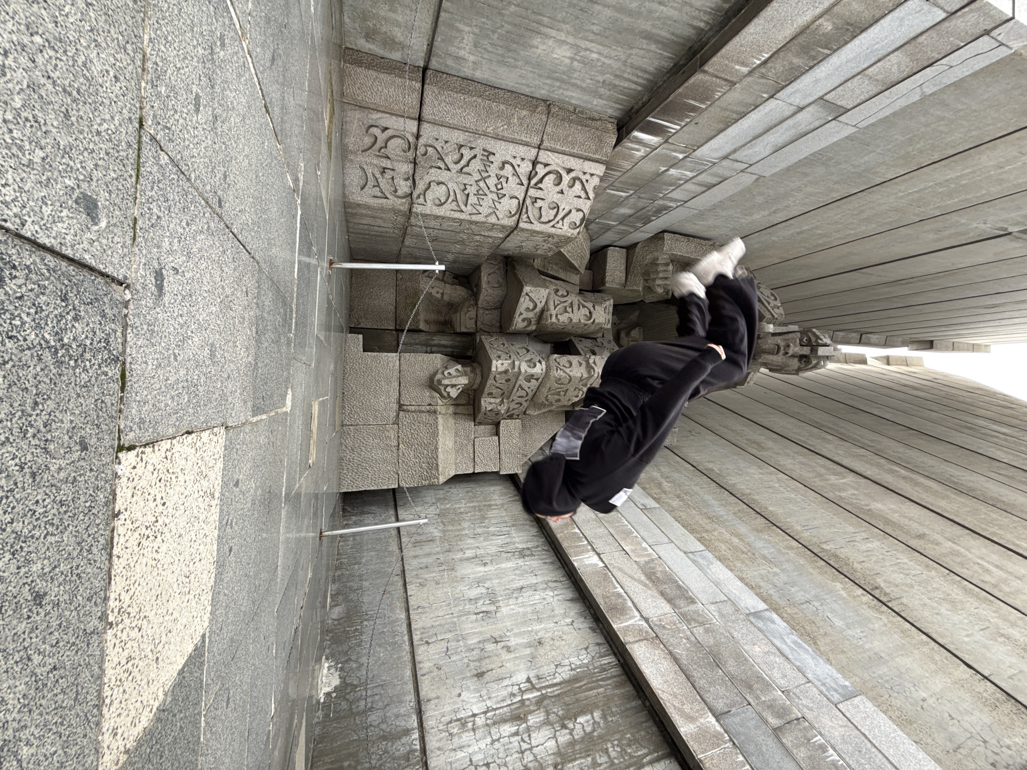 The image depicts a person in black clothing performing a handstand or acrobatic move at the Founders of the Bulgarian State Monument in Shumen, Bulgaria. The person's body is angled towards the right side of the frame, with their legs extended upwards and feet near the upper right corner. The monument's architectural details include textured stone surfaces, geometric patterns, and stylized ornamentation. Metal rods or cables are visible, possibly part of the monument's construction or support structure. The scene is composed of gray and off-white hues, with the architectural elements dominating the backdrop.