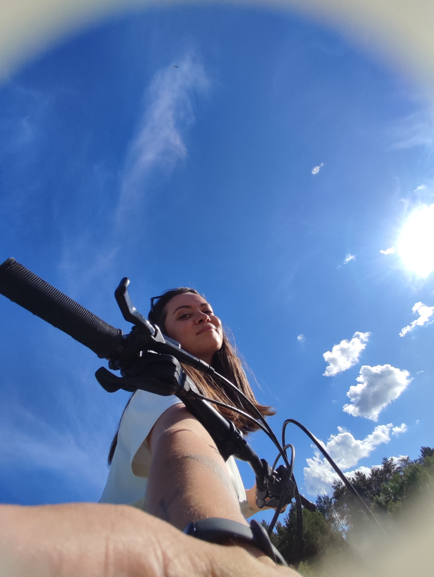 A low-angle, wide-angle shot captures a woman on a bicycle, under a bright blue sky dotted with clouds and the sun shining brightly. The woman has long brown hair, a white top, and is holding the handlebars of a black mountain bike. Part of her arm and a wrist-worn device are visible in the foreground. In the background, there is a treeline. High in the sky is what seems to be a airplane. The exact location in the city of Chaikovsky, Russia is not identifiable from this image.