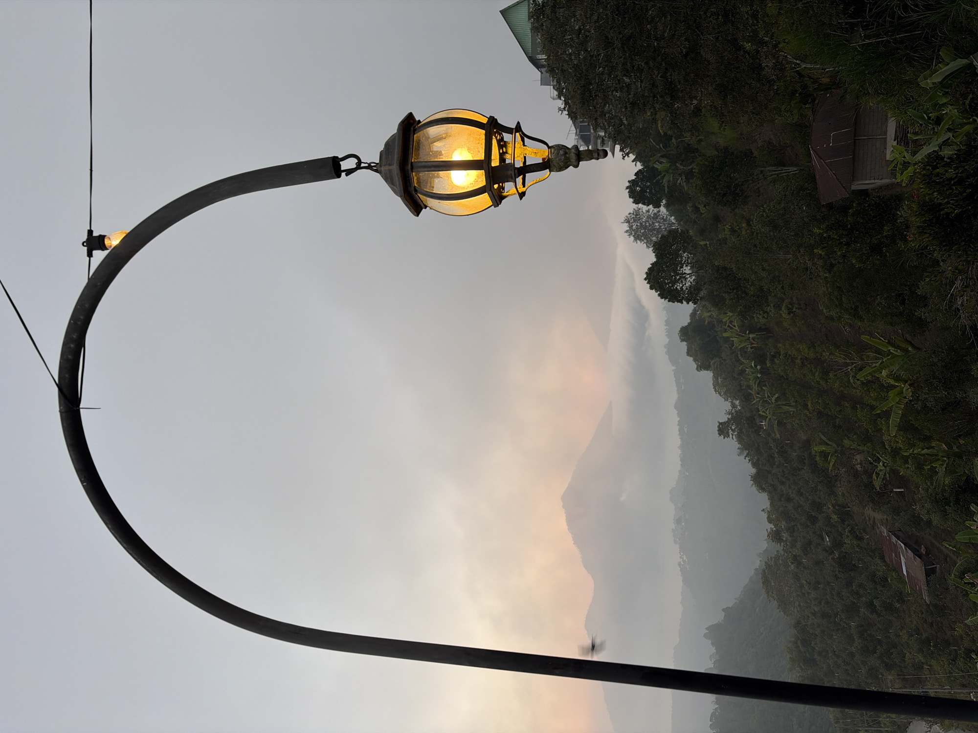 A lantern hanging from a curved metal pole is in the foreground of this image, with a misty mountain landscape in the background. The lantern, made of dark metal and glass, glows with a warm yellow light. Behind it, mountains rise through a thick layer of fog, with the faint glow of sunrise visible behind them. The foreground features lush green vegetation and trees, and a small, rustic building with a corrugated metal roof is nestled on a slope. The overall scene suggests a peaceful, early morning in a mountainous region, possibly Kintamani, Indonesia, known for its scenic beauty and volcanic landscapes.