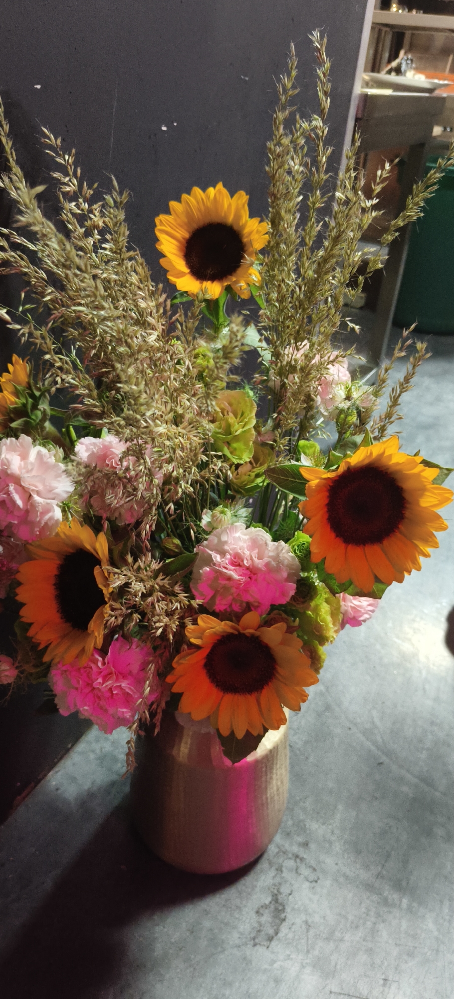 A bouquet of sunflowers and carnations is displayed in a metallic vase against a dark, textured wall. The sunflowers, with their vibrant yellow petals and dark centers, are the most prominent flowers. They are interspersed with delicate pink carnations and feathery, dried grass. The lighting in the scene is dramatic, casting shadows and highlighting the textures of the flowers and the vase. The background wall is a deep, charcoal gray, with subtle variations in tone. The floor is a concrete-like surface with a light gray hue and some darker, irregular marks. The overall impression is one of natural beauty captured in a somewhat moody and artistic setting. The location is not specified beyond "Unknown, Unknown."
