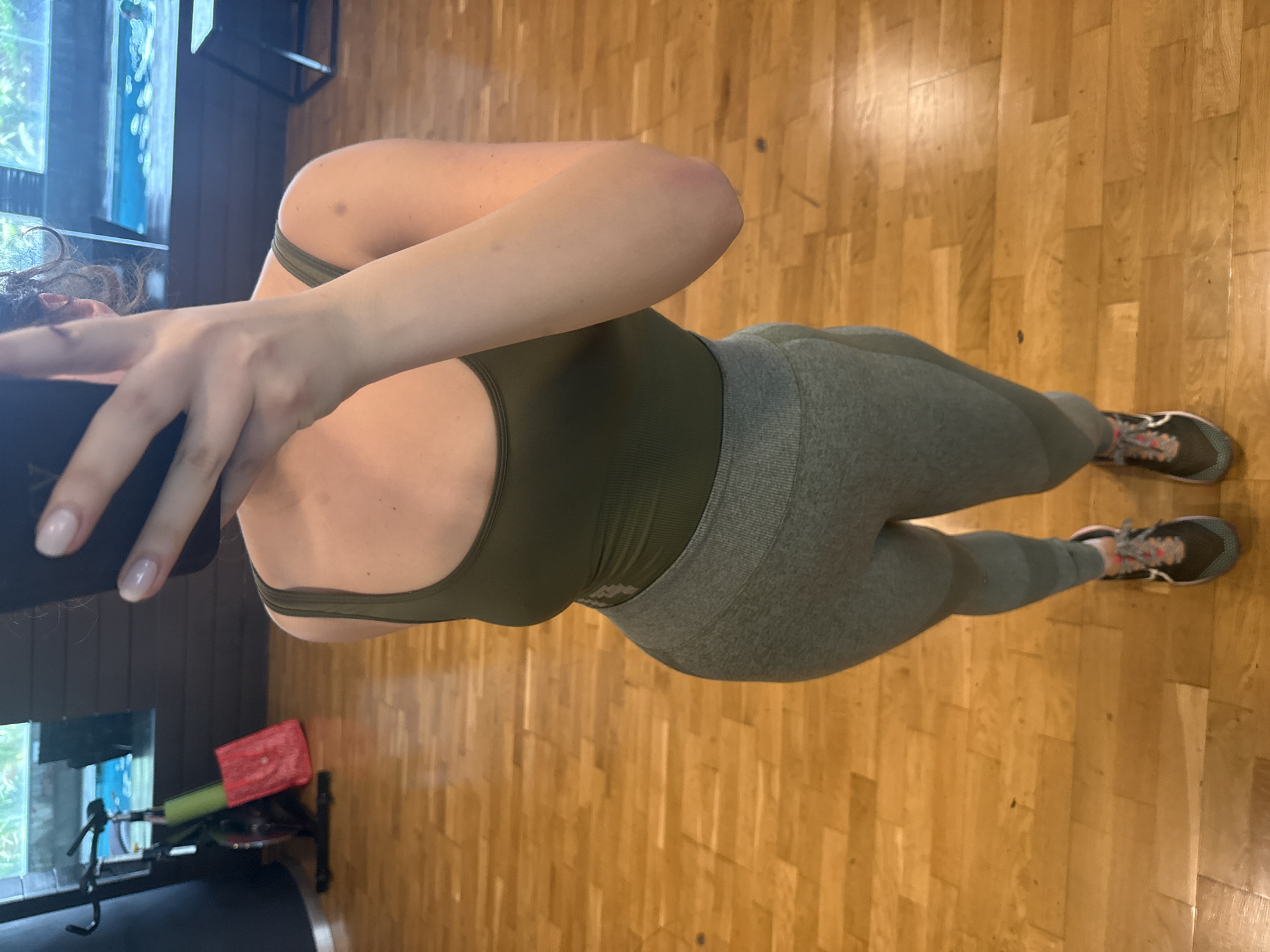 The image shows a woman in athletic wear, specifically a green sports bra and matching leggings, standing on a wooden floor. Her face is partially obscured by her hand holding a phone or similar device. The floor is made of light-colored wooden planks, and the background includes a window, exercise equipment (including a stationary bike), and other gym equipment. The location is possibly in Mexico City, Mexico.